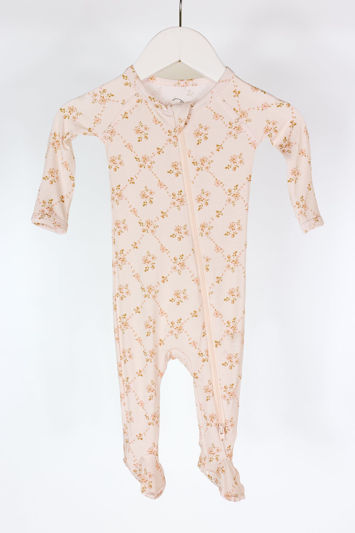 A light pink keepsake one piece zipper bamboo baby sleeper with long sleeves and feet, featuring a delicate floral pattern and a diagonal zipper, hangs on a white hanger against a plain white background.