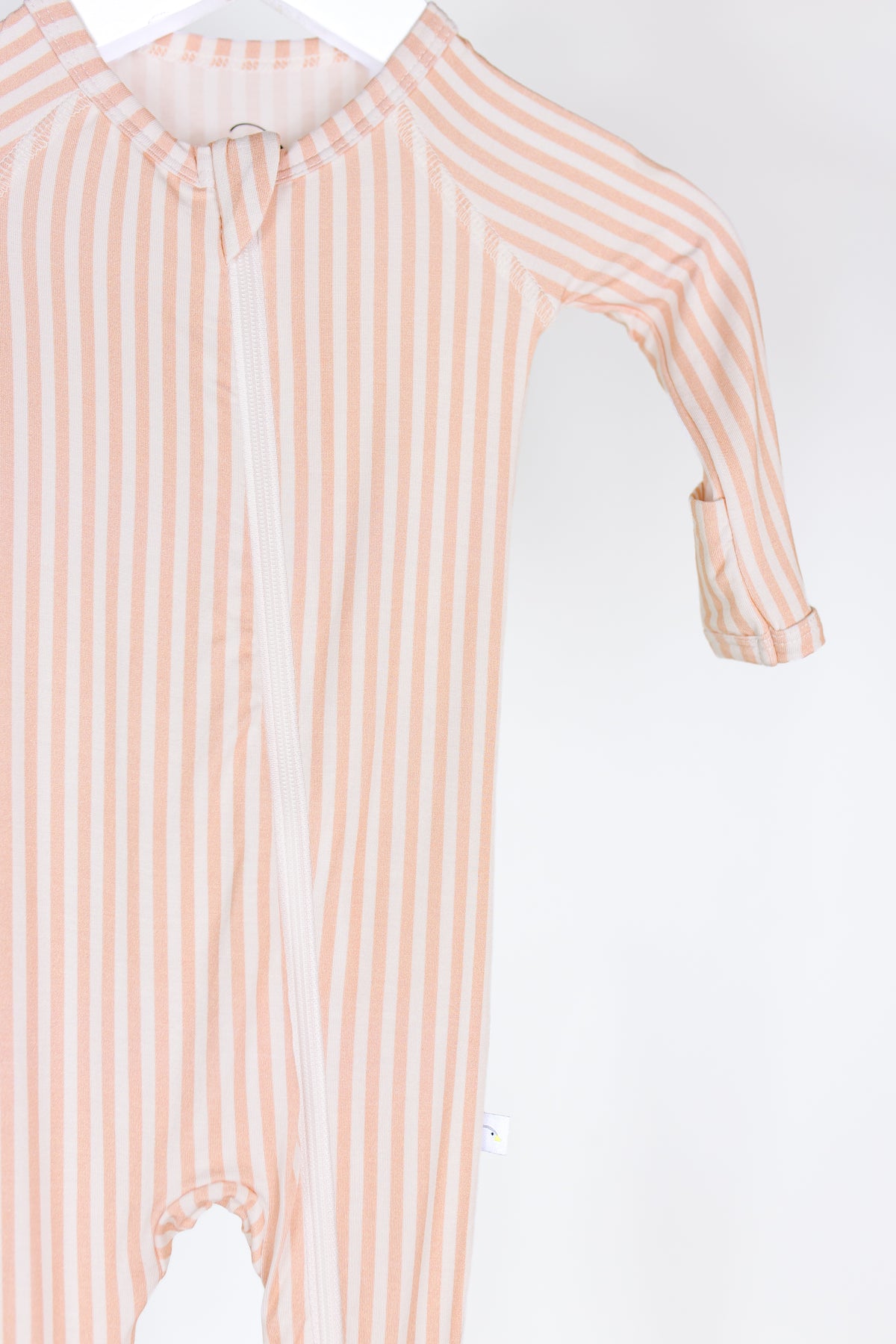 The heritage / one piece zipper is a long-sleeved baby onesie with light pink and white vertical stripes, hanging on a white hanger against a plain white background and featuring a convenient two-way zipper for easy changes.