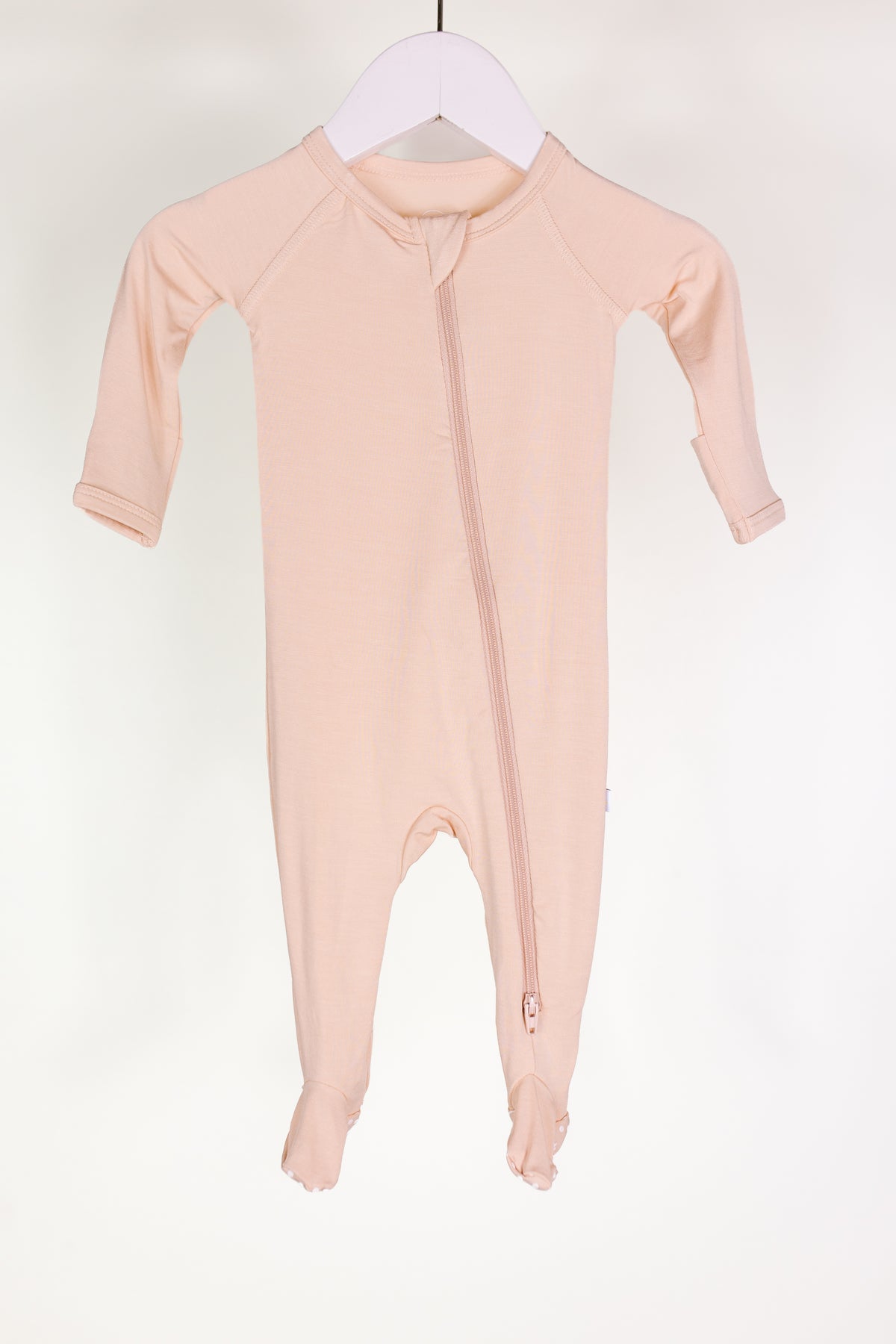 A whisp one piece zipper, a light pink bamboo baby pajama with long sleeves and covered feet, hangs on a white hanger. This newborn onesie features a diagonal front zipper running from the neck to the left ankle. The background is plain white.