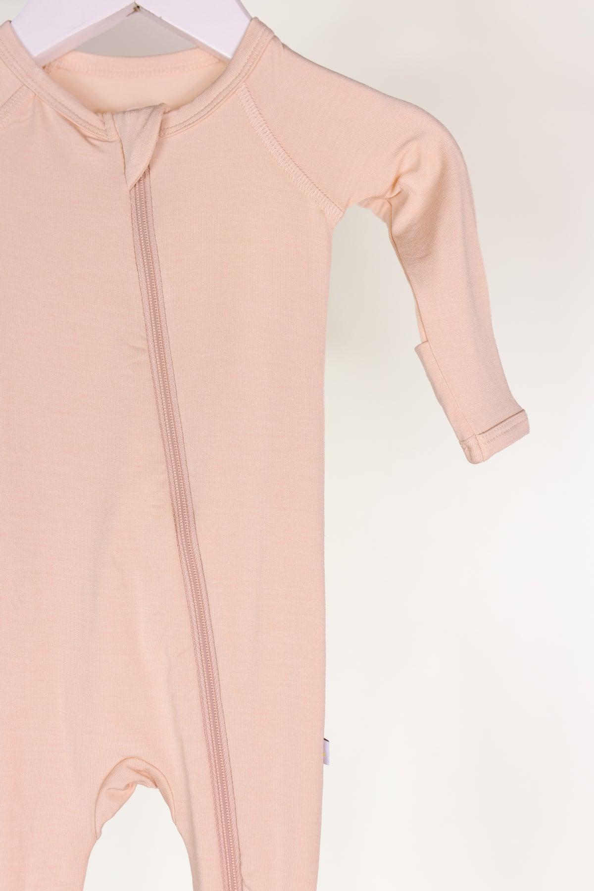 The whisp one piece zipper is a pale pink long-sleeve newborn onesie featuring a diagonal zipper, shown hanging on a white hanger against a plain white background.