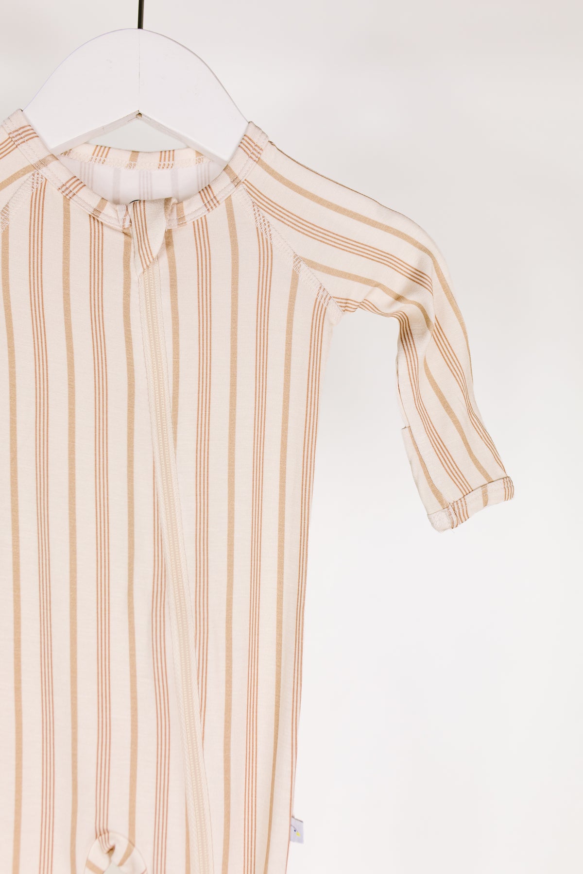 A “toast and honey / one piece zipper” bamboo baby pajama onesie in beige with vertical brown stripes is hanging on a white hanger against a plain white background, showing only the upper part and one sleeve.