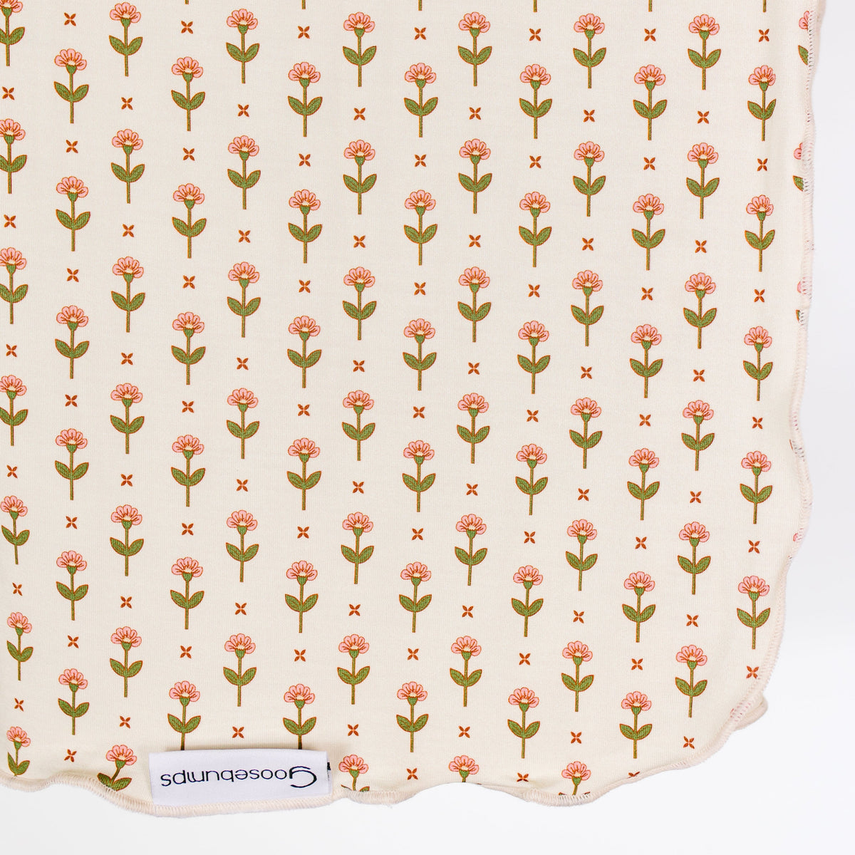Introducing the “tulip / swaddle”: a cream-colored bamboo baby blanket featuring a repeating pattern of green and pink tulips, each flower playfully separated by a small orange X. Its wavy edge and visible goosebumps tag at the bottom add distinctive charm to this single-layer swaddle—an irresistibly delightful choice for your little one.