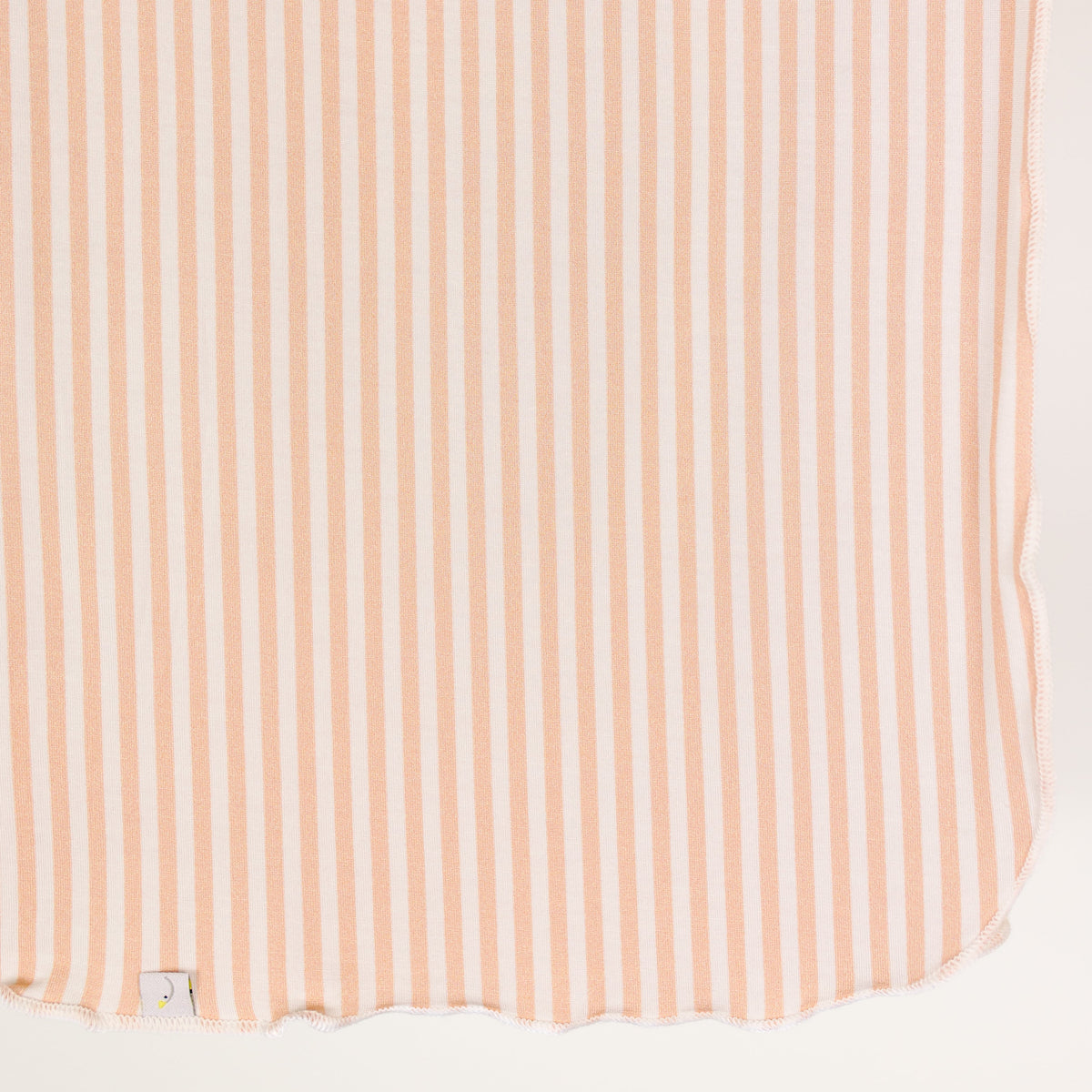 A heritage / swaddle featuring vertical light pink and white stripes, slightly curved edges, and a small white tag with a yellow dot in the bottom left corner.