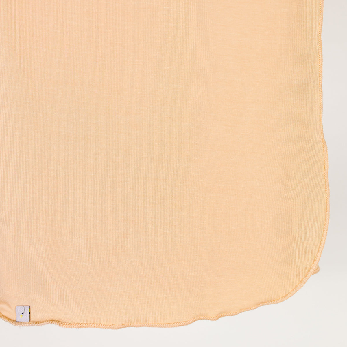 A close-up of the whisp / swaddle in a light peach hue, crafted from bamboo with stitched edges, gently curved at the bottom and featuring a small white tag near the lower left corner.
