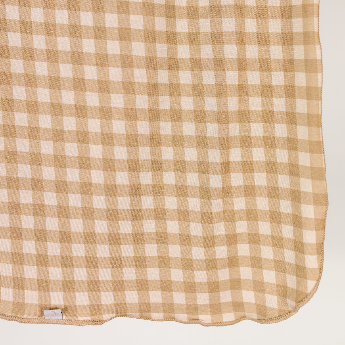 Introducing the teddy bear check / swaddle: a beige and white checkered fabric with a woven texture, neatly hemmed edges, and a small white fabric tag in one corner—this charming gingham swaddle blends timeless style with modern comfort.