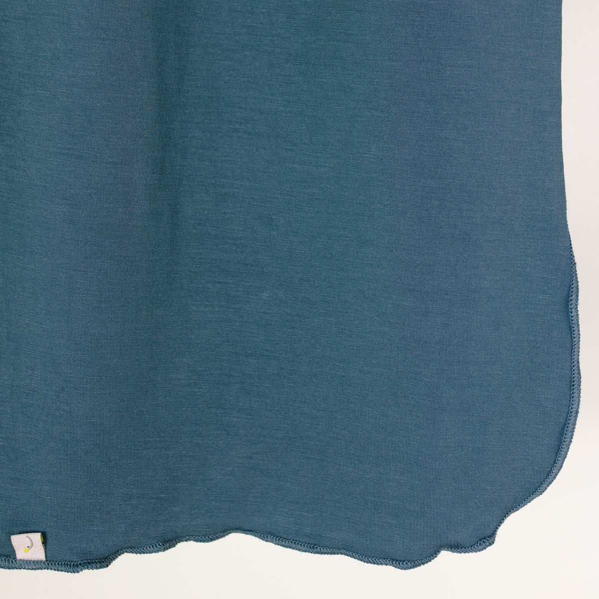 A close-up of the tidal wave / swaddle in indigo blue shows its stitched edges draped against a white background. Made as a single layer swaddle, the soft fabric displays a small tag in the lower left corner.