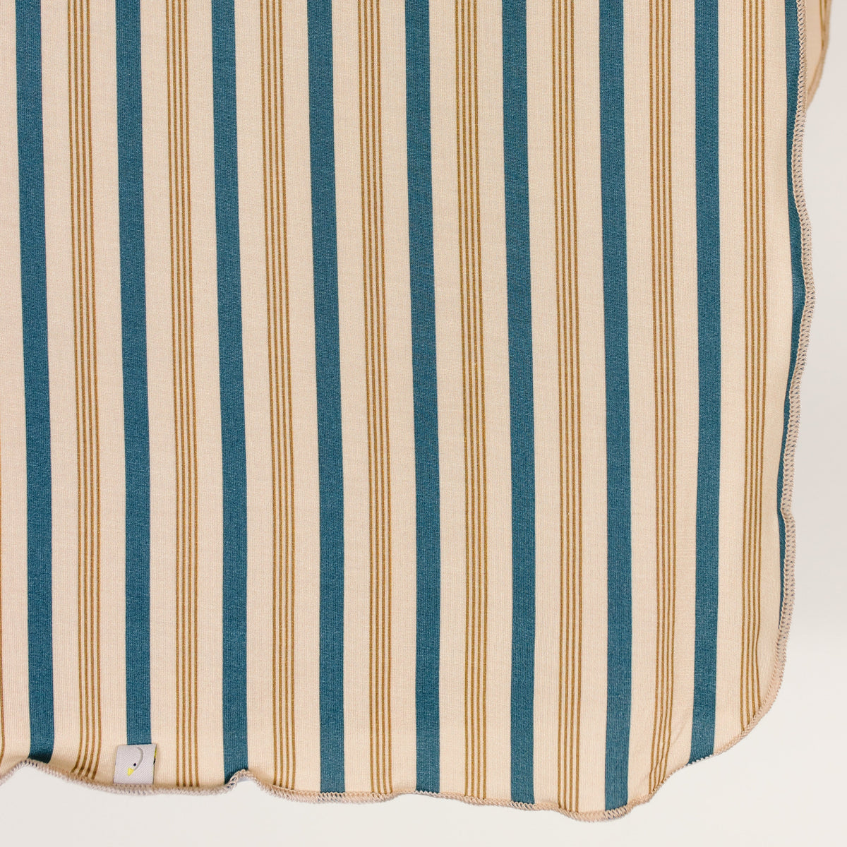 Crafted from soft beige fabric with evenly spaced vertical blue and tan stripes, the shoreline / swaddle features a wavy stitched edge and a small white tag in the bottom left corner—perfect for keeping your little one cozy.