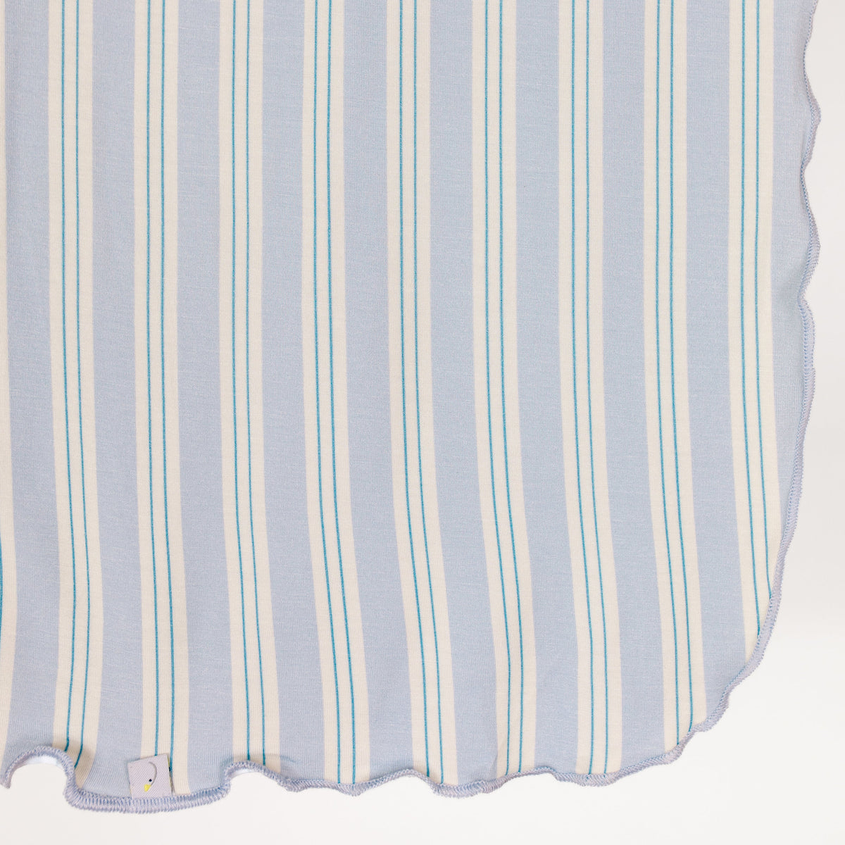 A light blue haven / swaddle from the Sarah Sweeten collection, crafted from bamboo fabric and adorned with vertical white and teal stripes, hangs with a gently scalloped edge. A small white tag is sewn into the lower left corner against a white background.