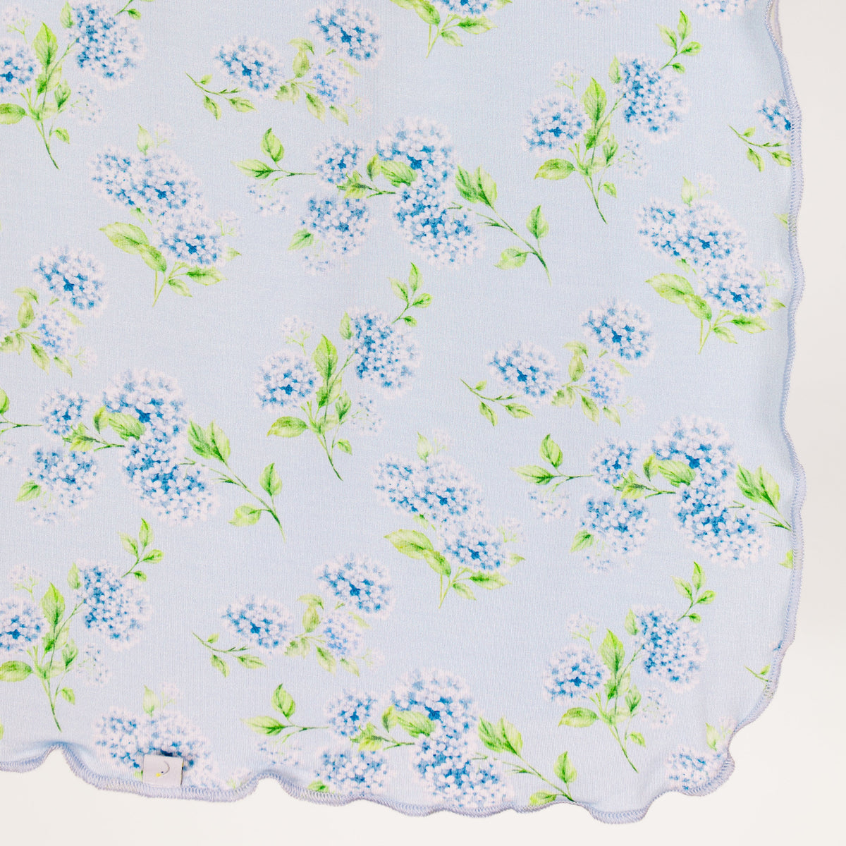 A light blue hydrangea swaddle from the Sarah Sweeten Goosebumps Collection, featuring a delicate pattern of blue and white lilac flowers with green leaves and finished with an elegant wavy, scalloped trim along the edge.