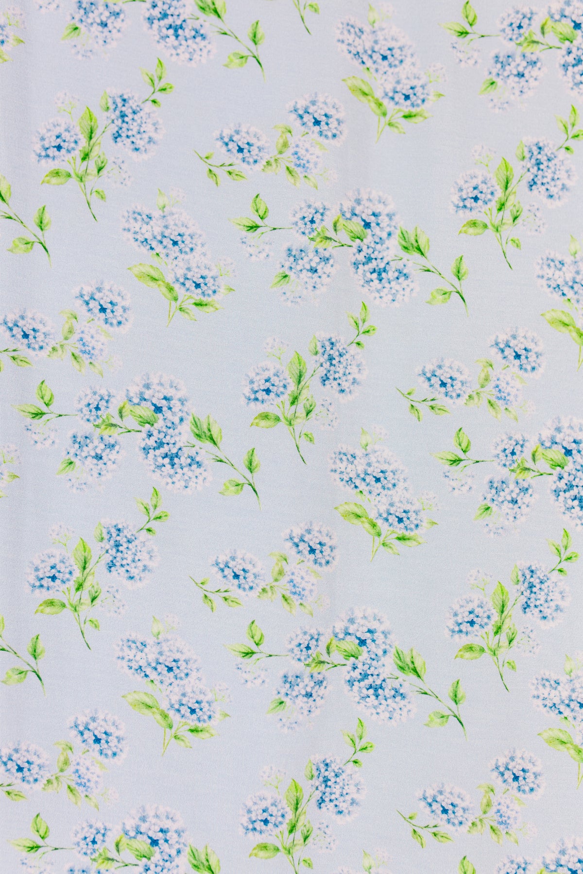 An evenly spaced pattern of small blue and white flowers with green leaves graces a light background, capturing the delicate, spring-like look of the hydrangea / swaddle—perfect for a soft and charming bamboo wrap.