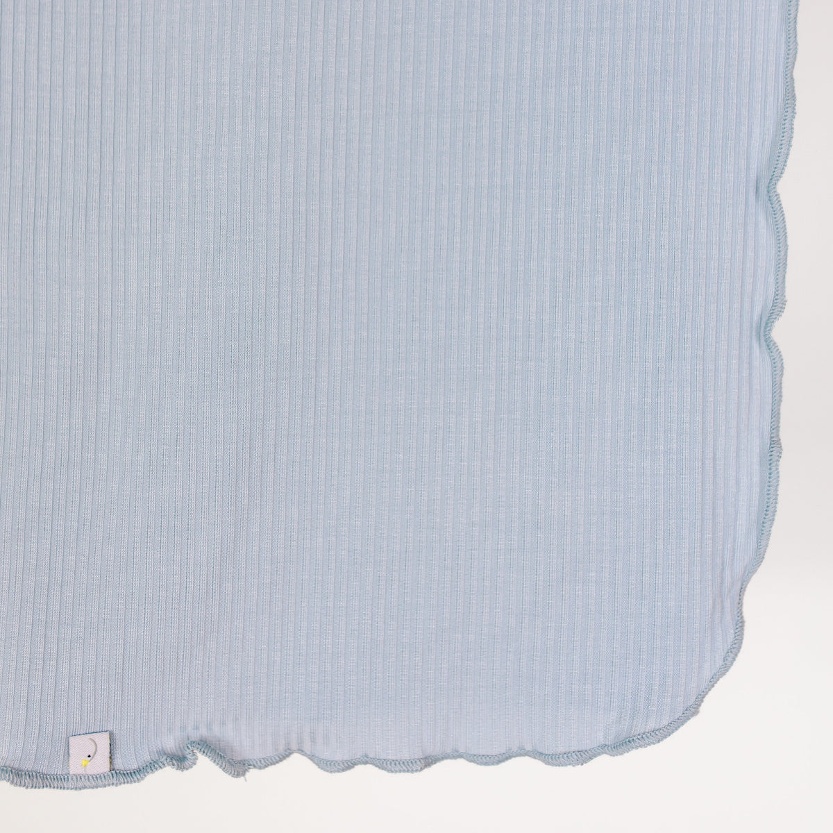 Featuring a light blue, ribbed fabric with wavy, scalloped edges, the cove / swaddle showcases a small white tag sewn into the lower left corner. The bamboo swaddle is set against a white background and occupies most of the image.