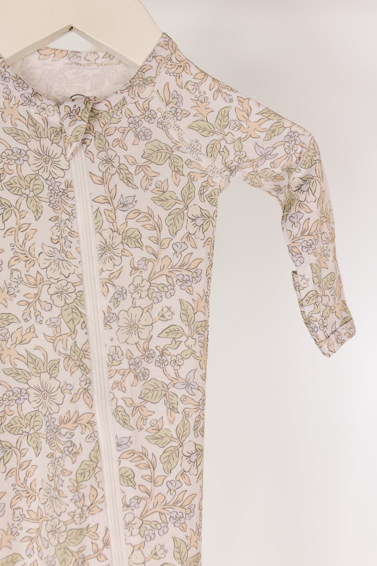 A close-up of the heirloom / one piece zipper—a bamboo newborn zipper onesie featuring a soft pastel floral pattern and a front zipper—hanging on a white hanger against a plain background.