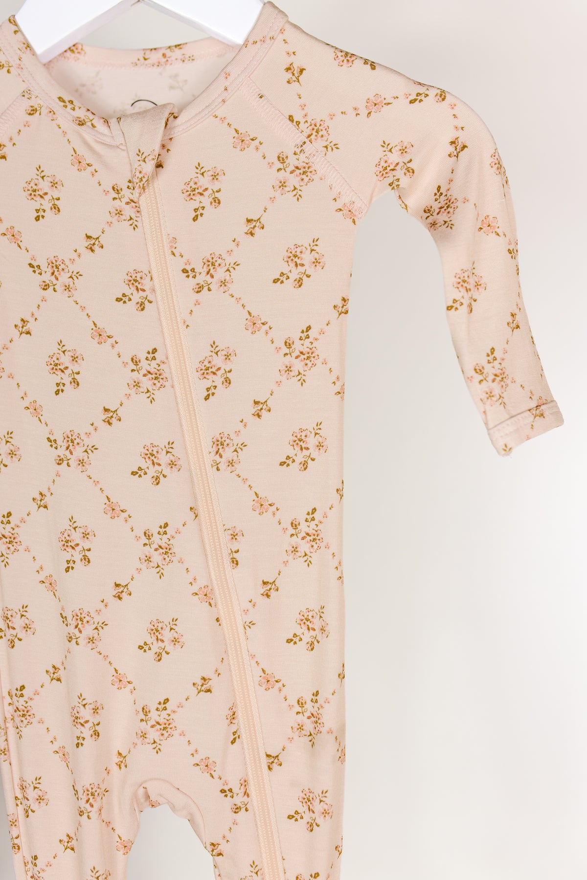 A pale pink “keepsake / one piece zipper” made from soft, breathable bamboo fabric, featuring a delicate floral pattern and a convenient front zipper, hangs on a white hanger against a neutral background.
