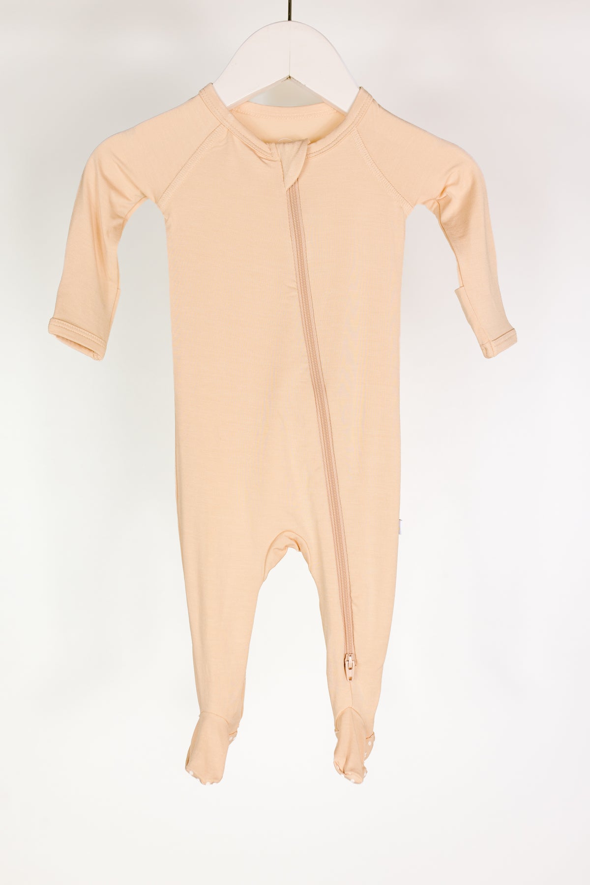 The whisp one piece zipper is a beige, long-sleeve bamboo baby pajama with footed bottoms, shown on a white hanger. These cozy pajamas feature a diagonal zipper from the neck to one leg, making changes easy. The background is plain white.