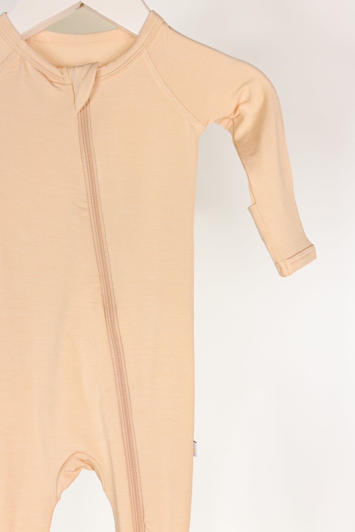 Whisp / one piece zipper in beige bamboo with long sleeves and a diagonal zipper, hanging on a white hanger against a plain white background. Only the upper part of this cozy newborn zipper onesie is visible.