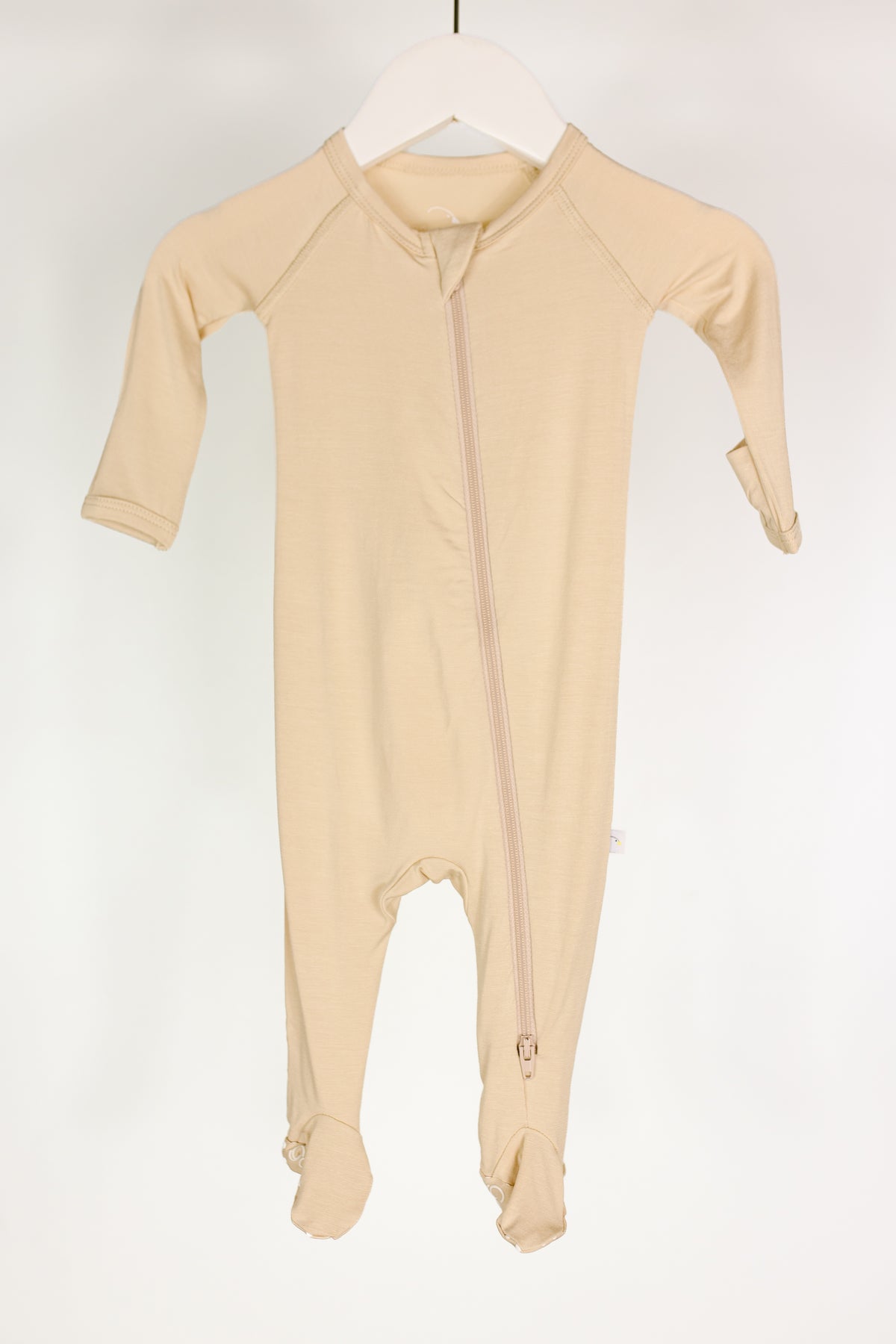 A vanilla cream one piece zipper newborn onesie with long sleeves and built-in footies hangs on a white hanger. The asymmetric zipper runs from the neckline down to the left ankle, making this cozy footed pajama both stylish and practical. The background is plain white.