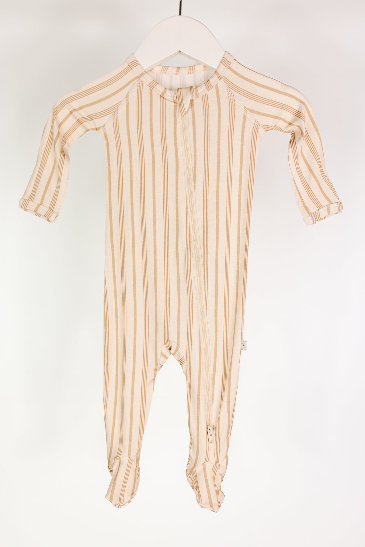 The toast and honey / one piece zipper is a cream-colored baby onesie with long sleeves and legs, adorned with thin tan vertical stripes. Hung on a white hanger against a plain white background, these cozy footed pajamas are designed for comfort and feature a practical two-way zipper to make diaper changes easy.