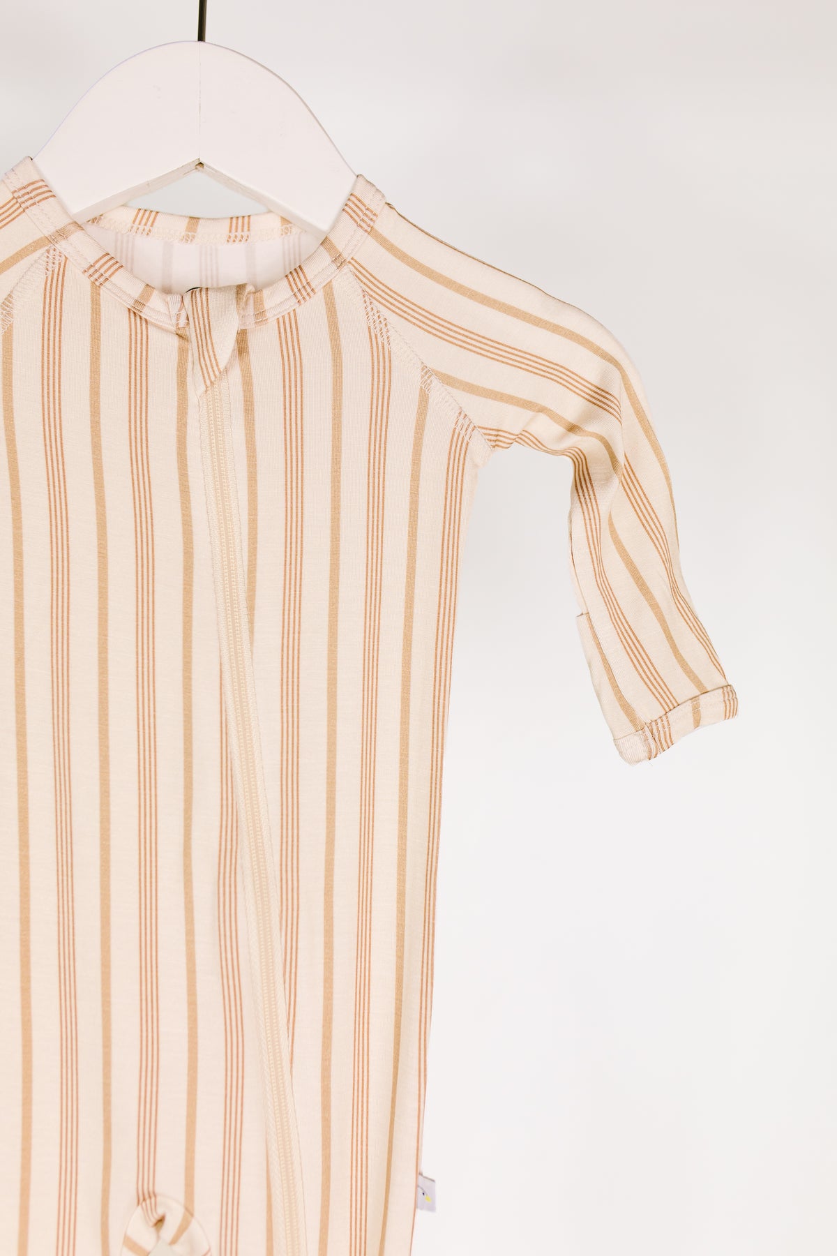 A toast and honey one piece zipper baby onesie with brown vertical stripes hangs on a white hanger against a plain white background. Showcasing the upper half and one sleeve, this cozy piece evokes the classic style of footed pajamas for newborns.