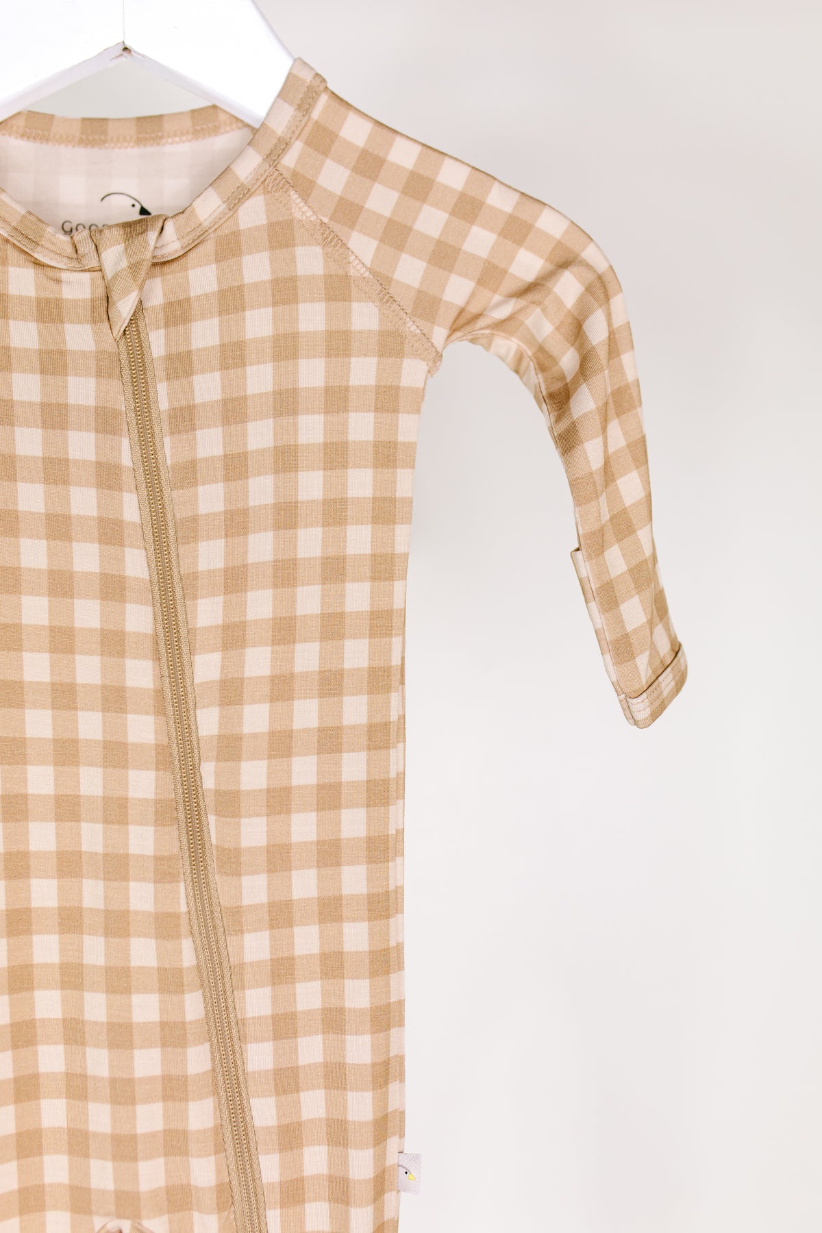 The teddy bear check one piece zipper, a beige and white plaid newborn onesie with a front zipper, is hanging on a white hanger against a plain light background.