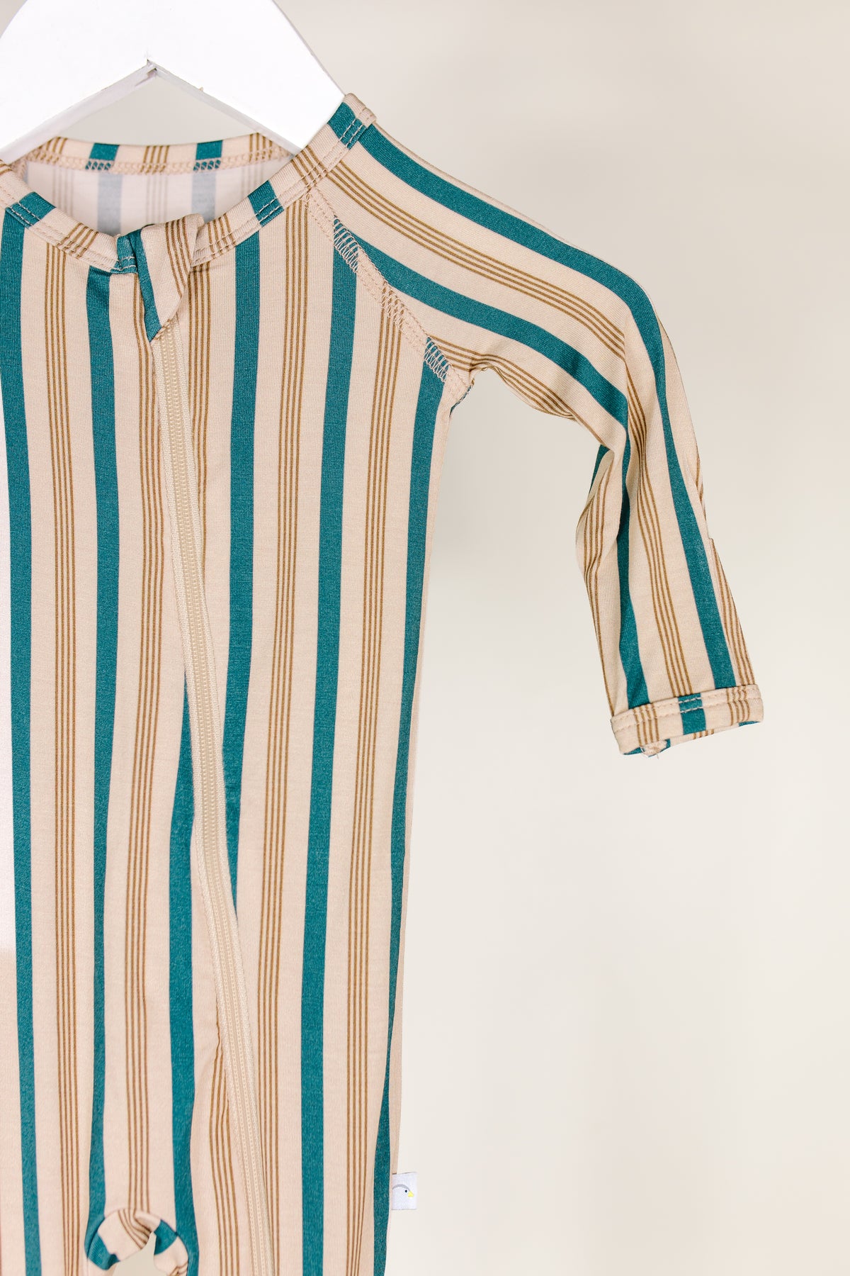 A close-up of the shoreline one piece zipper, a bamboo baby sleeper featuring teal and tan vertical stripes, displayed on a white hanger against a plain light background.