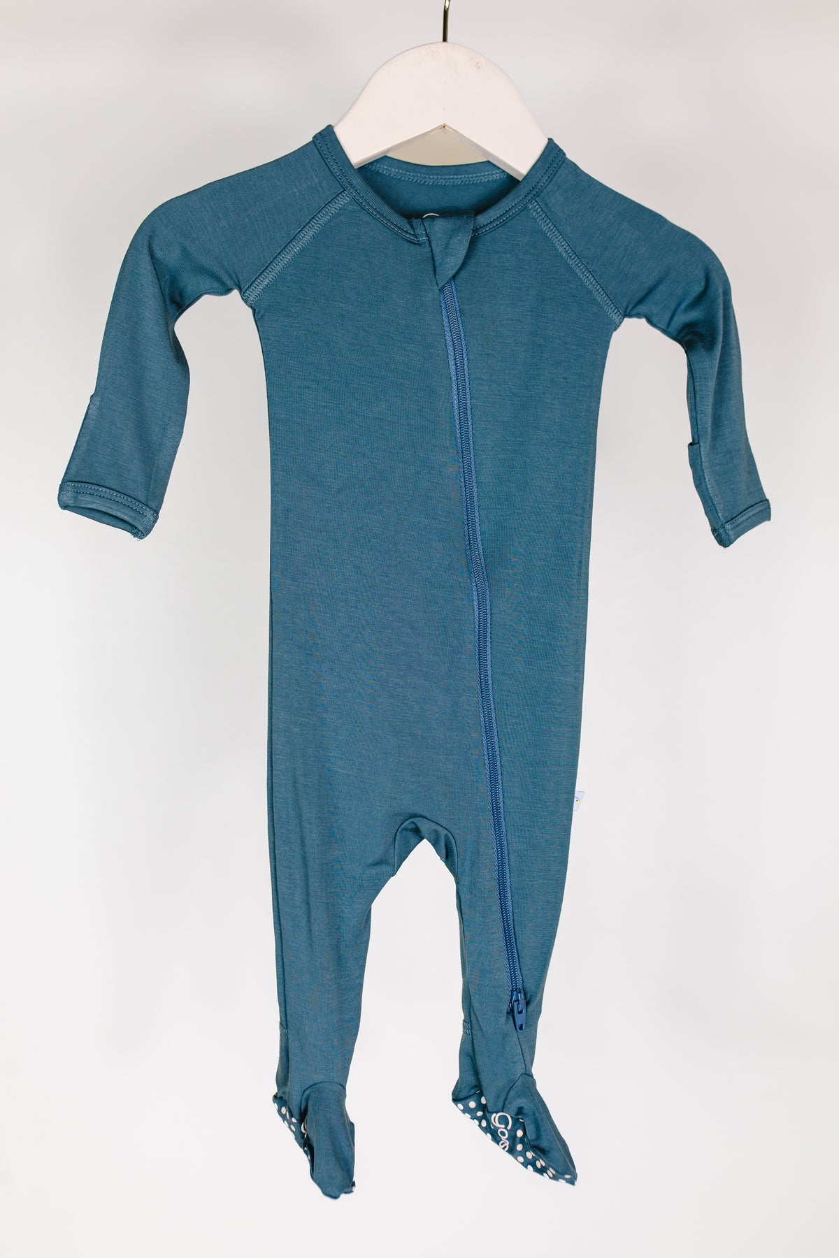 A tidal wave one piece zipper—a long-sleeve, teal blue bamboo baby pajamas onesie with covered feet and a diagonal front zipper—is displayed hanging on a white hanger against a plain white background.