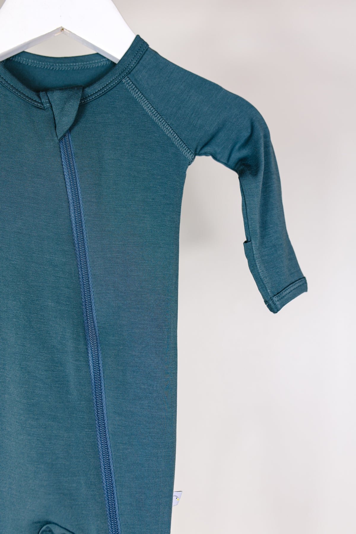 A close-up of the tidal wave one piece zipper, a teal long-sleeve bamboo baby pajamas onesie, hanging on a white hanger with one sleeve and part of the body visible against a plain background.