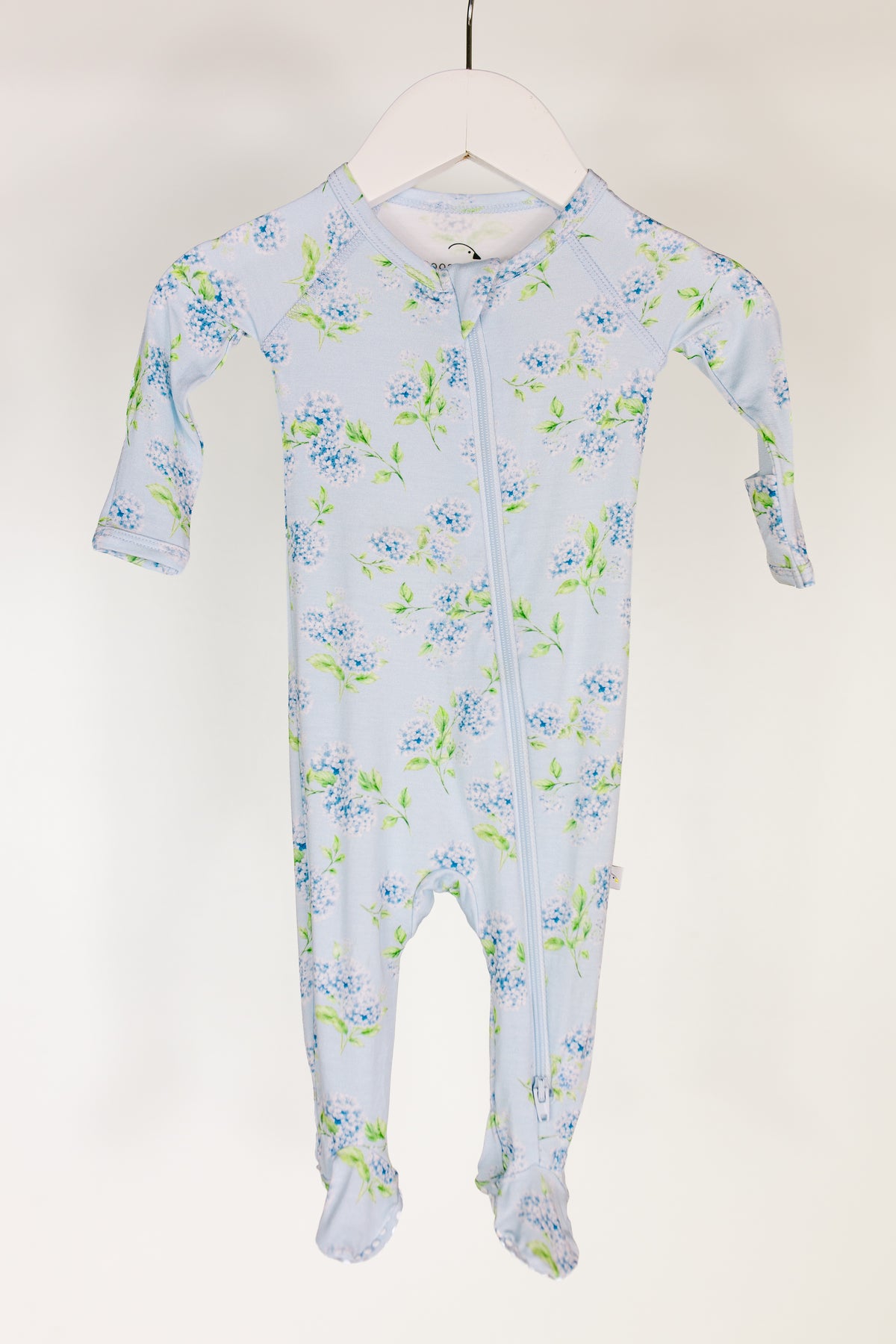 Dress your little one in the hydrangea one piece zipper, a light blue infant onesie adorned with a charming blue and green floral print. This buttery-soft newborn outfit features long sleeves, covered feet, a diagonal front zipper, and handy fold-over mittens for added comfort. The hydrangea one piece zipper is displayed on a white hanger against a simple background.
