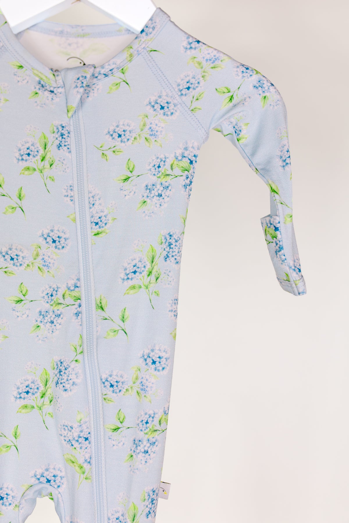 A hydrangea / one piece zipper baby onesie in light blue, featuring a hydrangea floral pattern and crafted from buttery-soft fabric, is displayed on a white hanger against a plain background. Only one arm and part of the leg are fully visible.