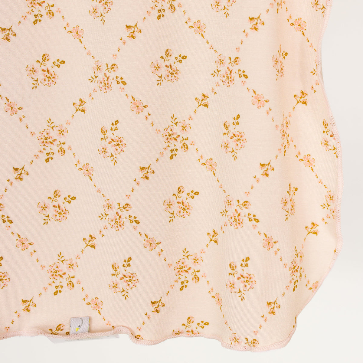 The keepsake / swaddle features a soft pink bamboo fabric adorned with a repeating diamond pattern of tiny flowers and leaves in light pink and gold tones. This delicate floral print is finished with a small fabric tag at the lower edge.