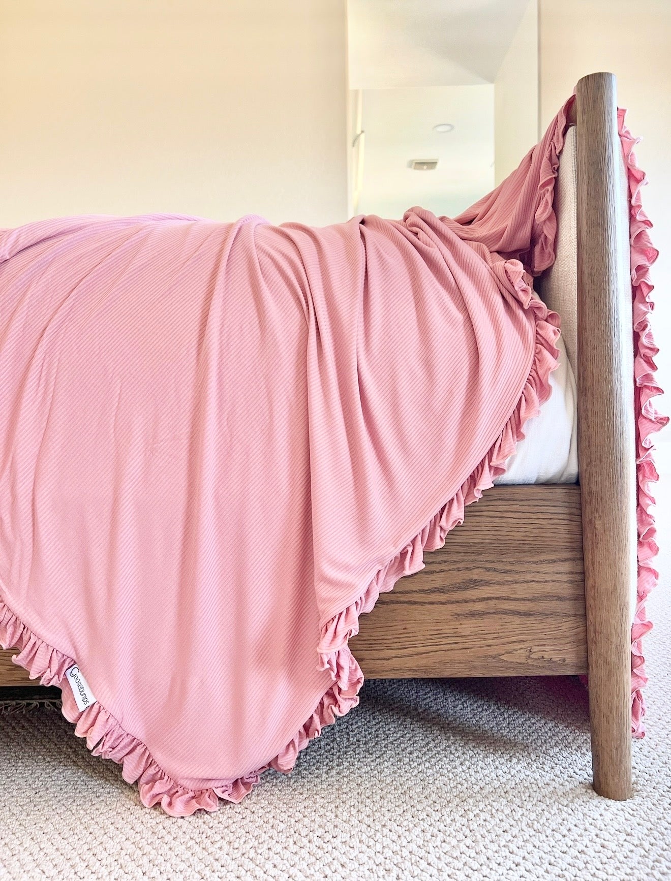 A minimalist, bright bedroom features a wooden bed adorned with the "peony / ruffle" luxurious pink blanket over crisp white bedding, set atop a light-colored carpet.