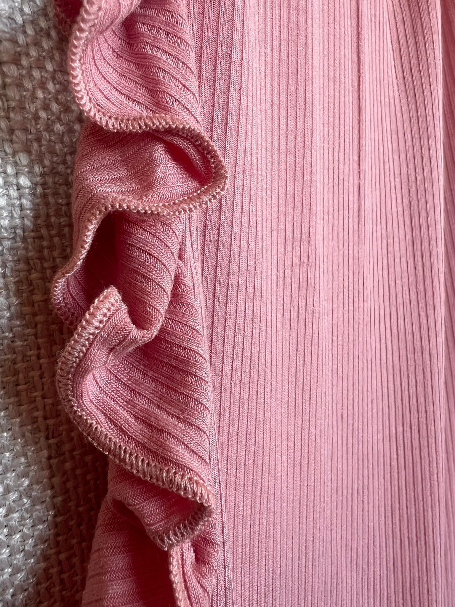 Close-up of the peony / ruffle blanket in ribbed pink with a wavy, ruffled edge beside textured off-white fabric. The decorative border along the seam highlights the elegant detail of this luxurious blanket.
