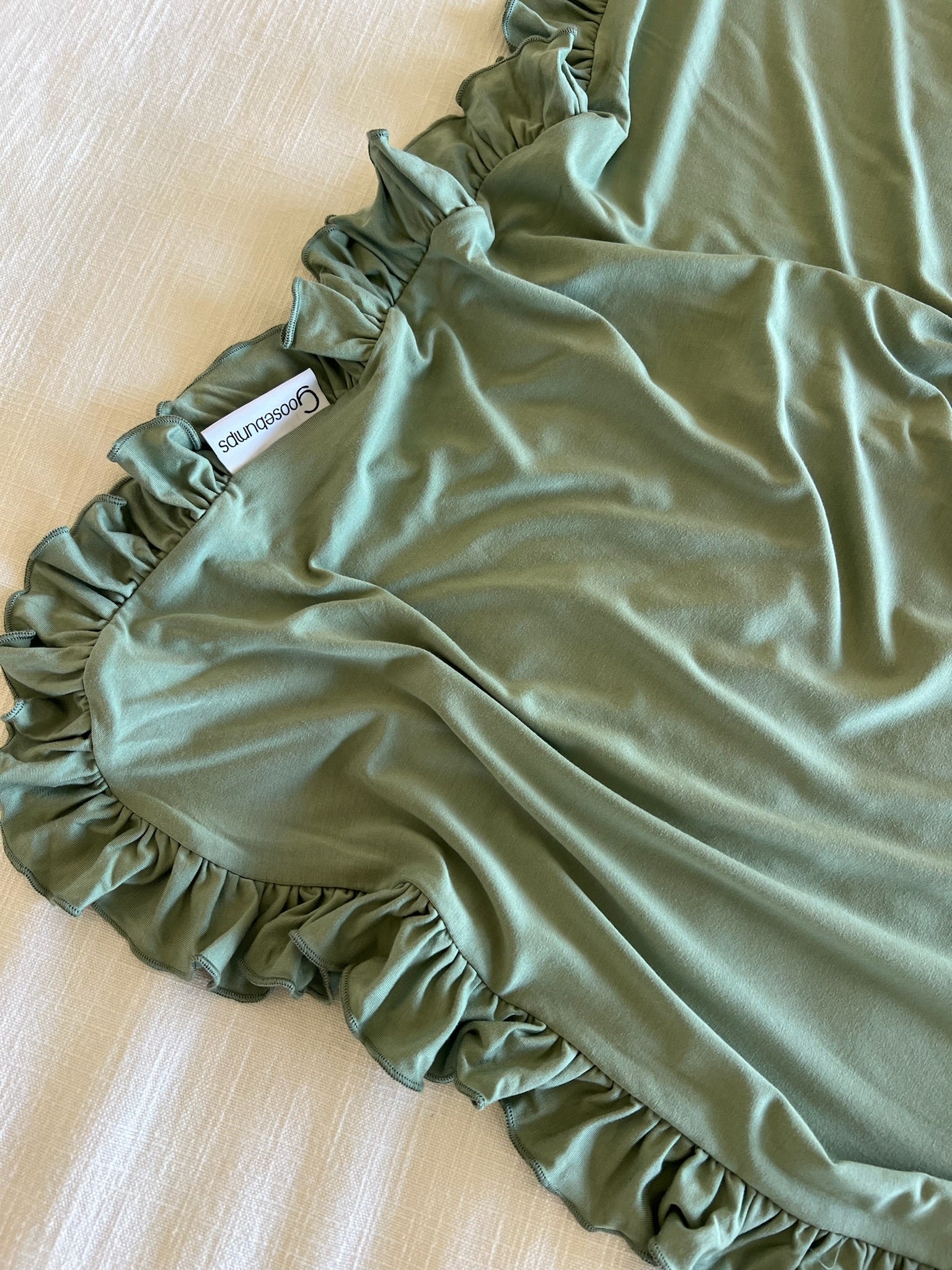 A soft, light green basil / ruffle blanket with delicate edges is spread out on a cream-colored textured surface. A visible clothing label reads SKIMS COTTON SLEEP.