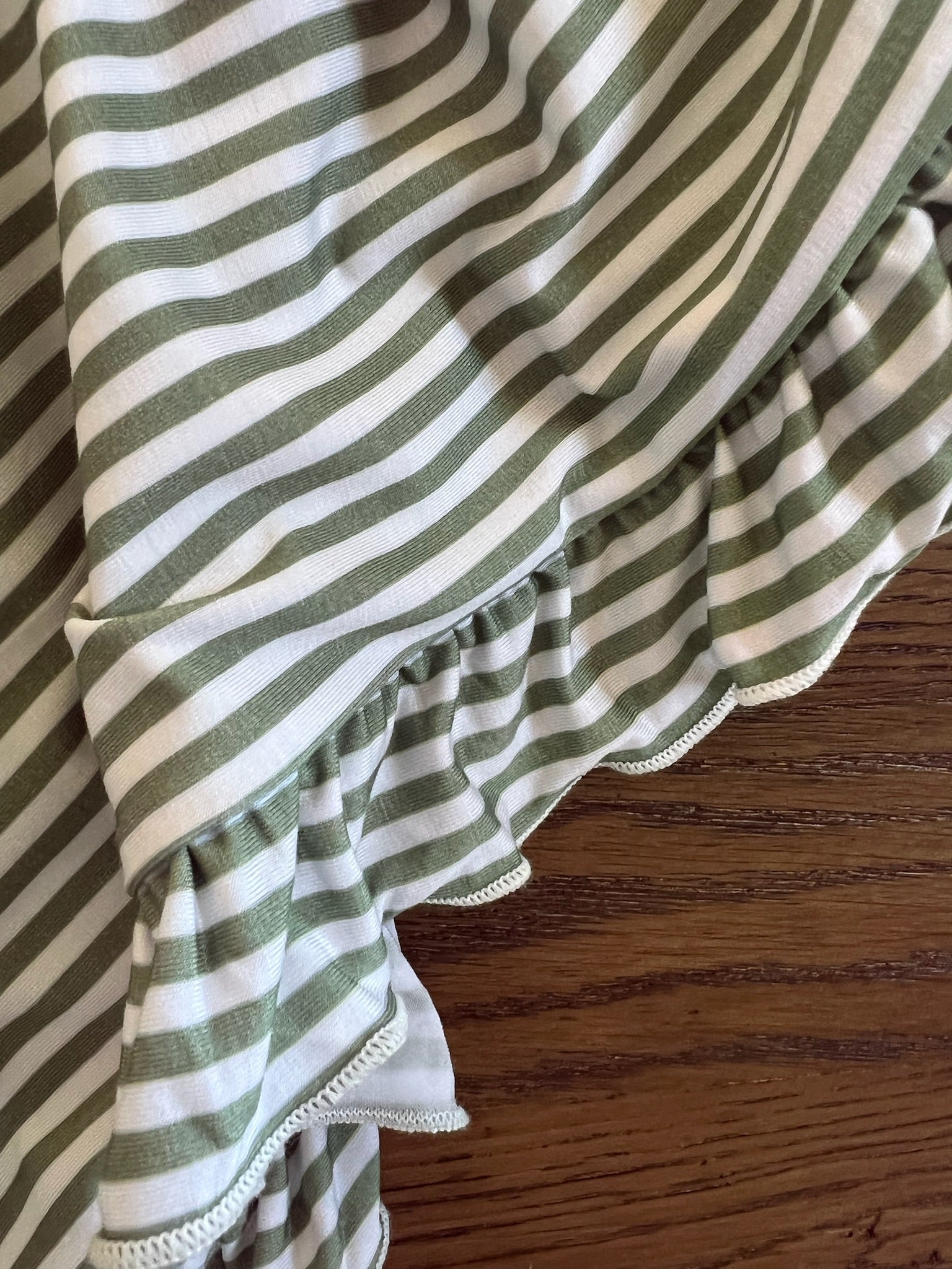 A close-up of the "tree house / ruffle" vintage striped blanket, featuring a ruffled edge and lying on a wooden surface with visible wood grain beside its soft, flowing fabric.