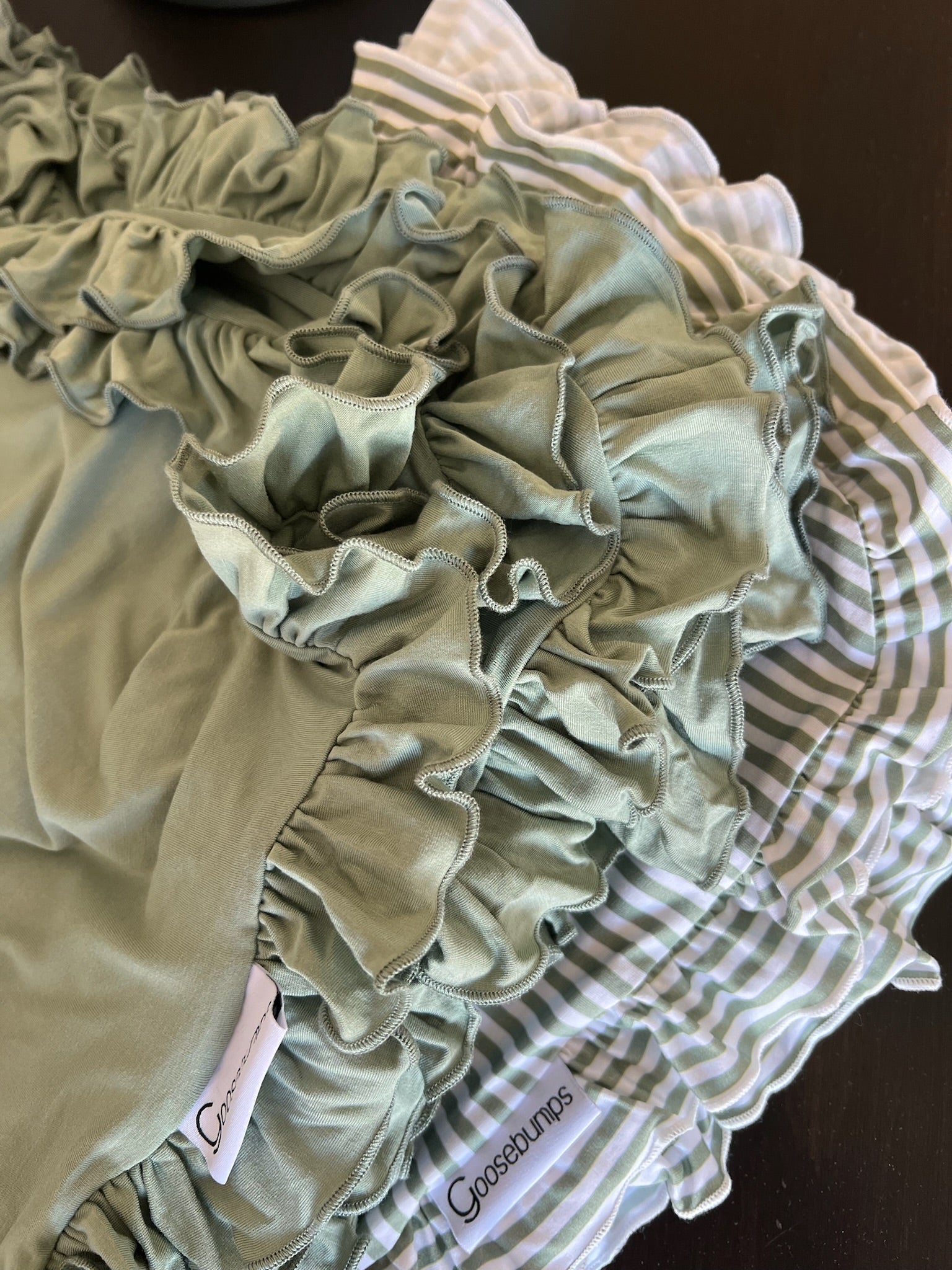 Two “tree house / ruffle” throws—one pale green, one green-and-white striped—overlap on a dark surface. Goosebumps tags are visible by the ruffled edges.