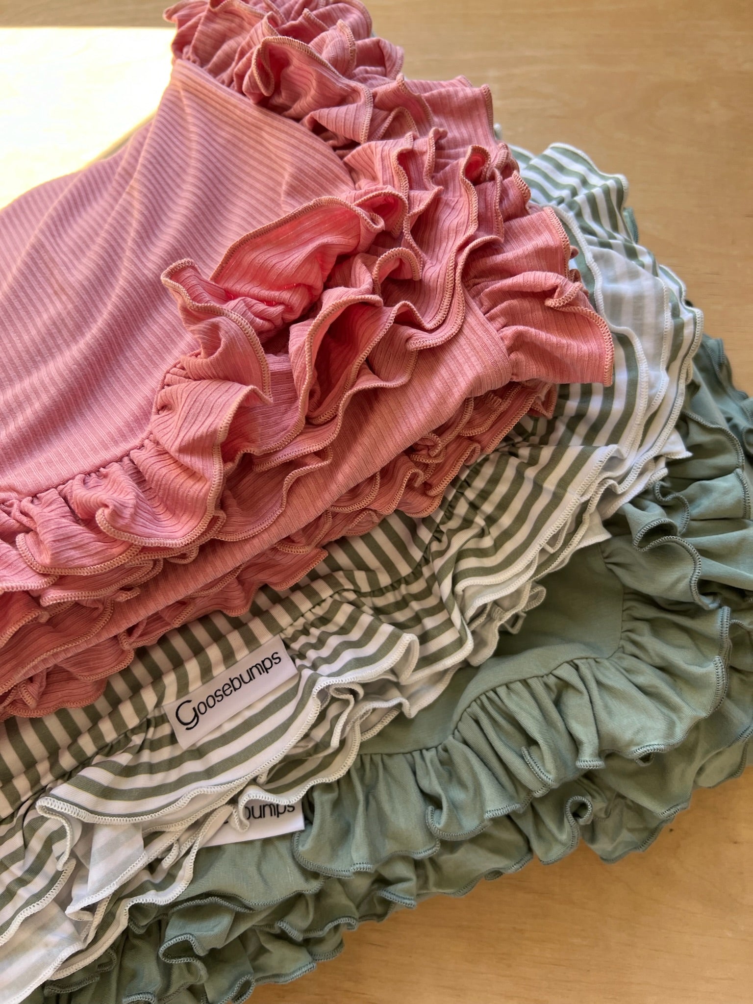 A stack of peony / ruffle baby bloomers in pink, green, and white stripes sits in sunlight on a wooden surface beside a soft pink ruffle blanket, adding a cozy touch.