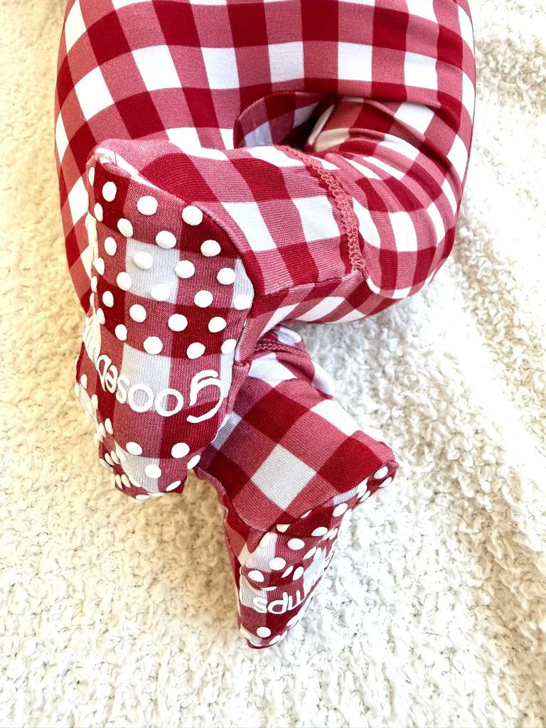 Newborn feet in red and white gingham checkin’ it twice / one piece zipper pajamas, featuring white non-slip grips on the soles.