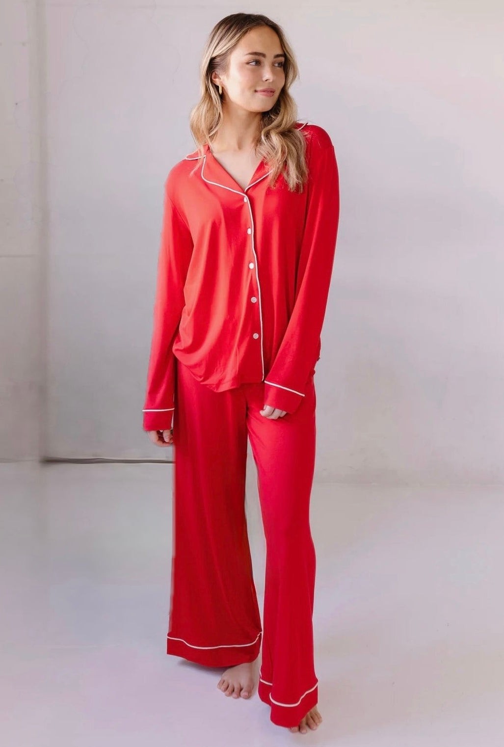 A woman stands barefoot indoors in a relaxed fit classic red pajama set with white piping, looking to the side against a plain, light-colored background.