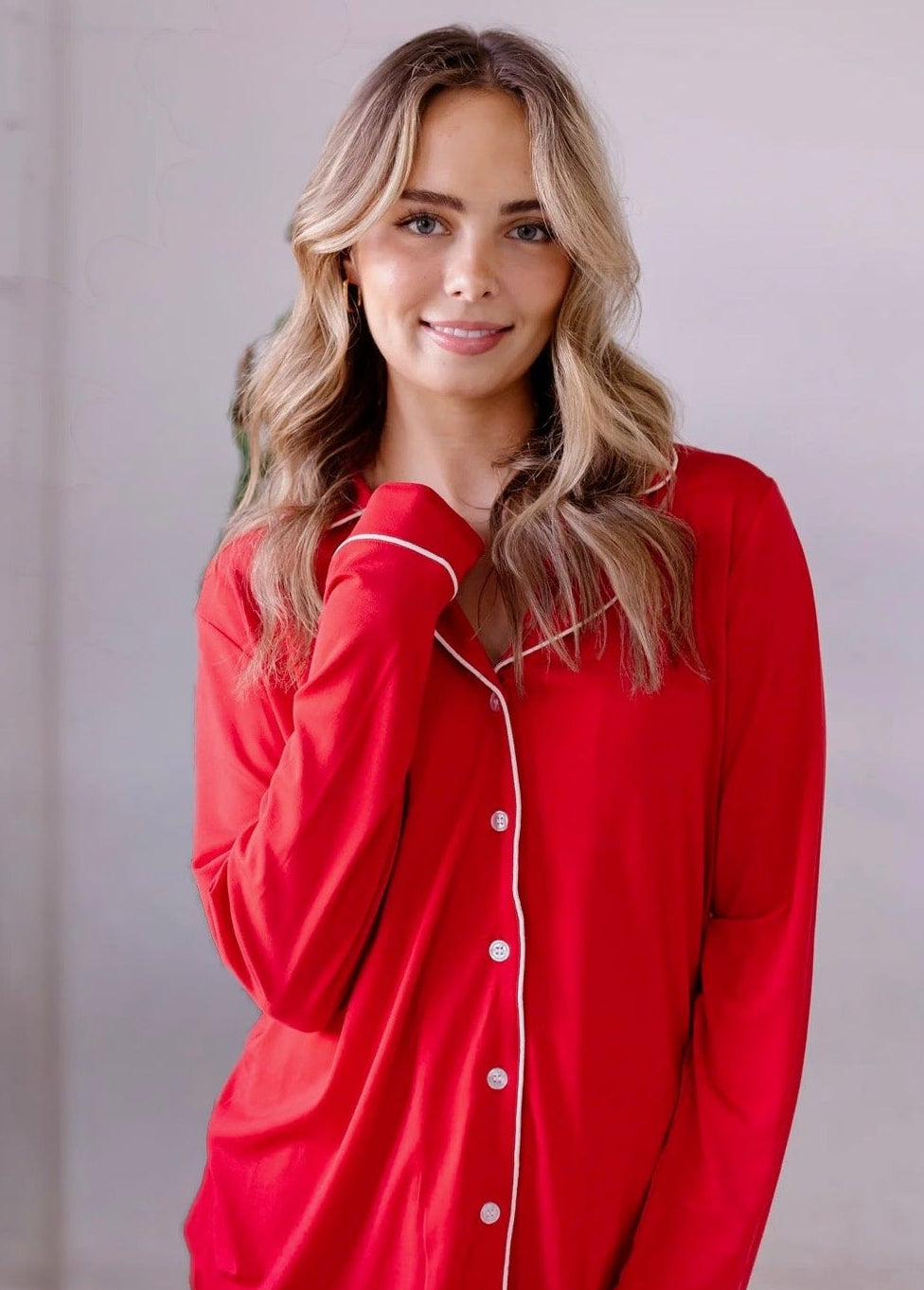 A smiling woman with wavy blonde hair wears the classic red/pajama set—relaxed fit bamboo viscose loungewear with white piping—from the holiday collection, standing indoors against a light background.