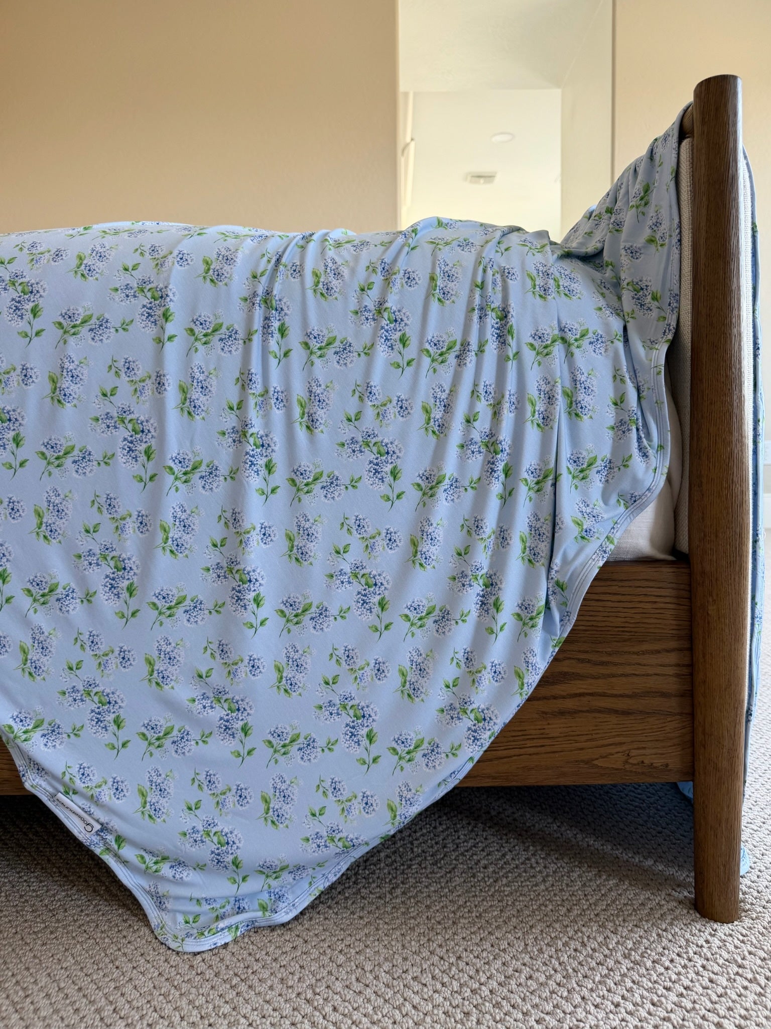 A wooden bed with light blue floral hydrangea sheets draped over the side sits against a beige wall and a light carpeted floor. Sunlight brightens the room, while a soft bamboo blanket from the Goosebumps Collection adds an extra layer of comfort.