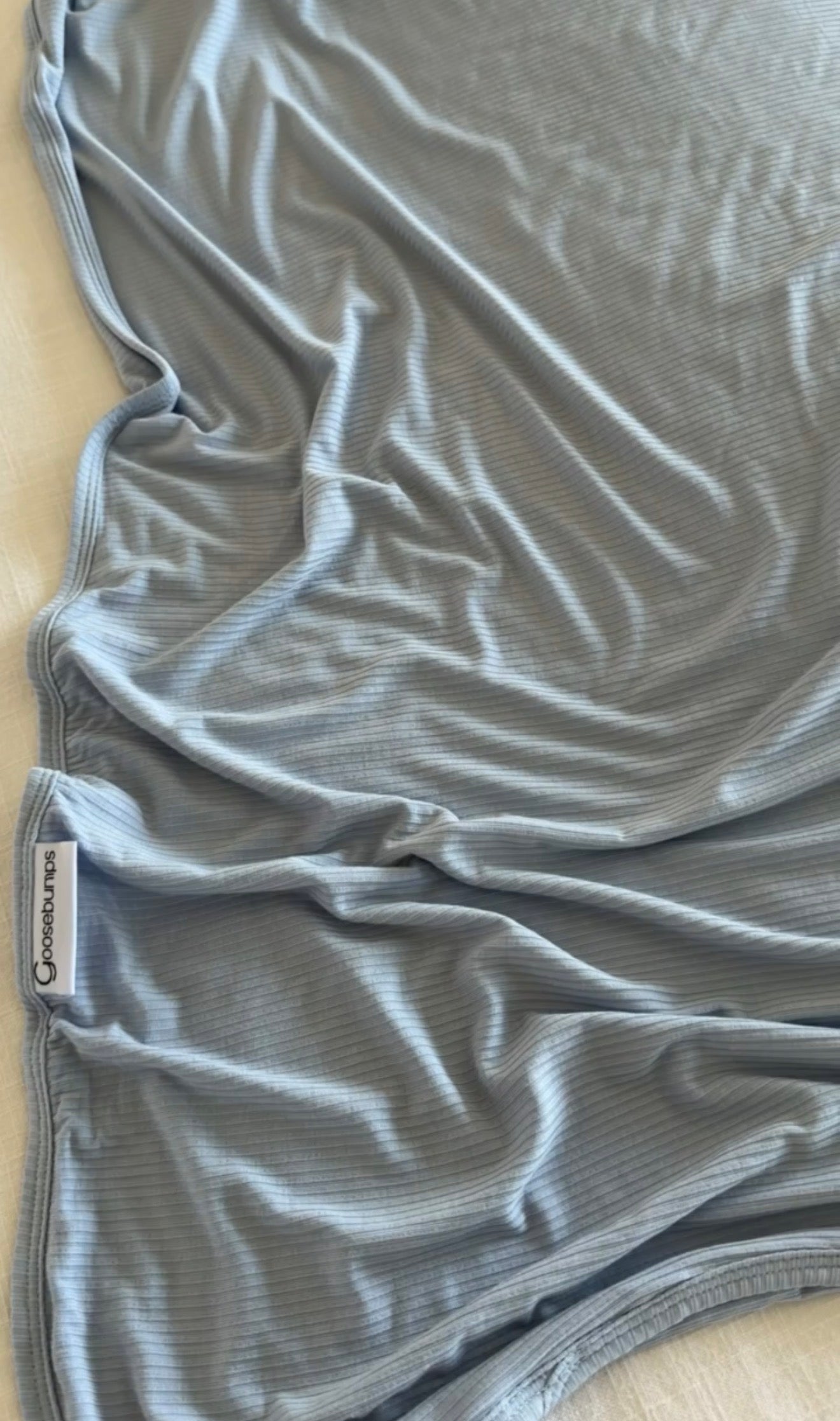 A luxurious light blue “Cove” blanket made from ribbed fabric with visible wrinkles is spread out on a surface. A small white tag with black text reading Copper Pearl is sewn into one edge.