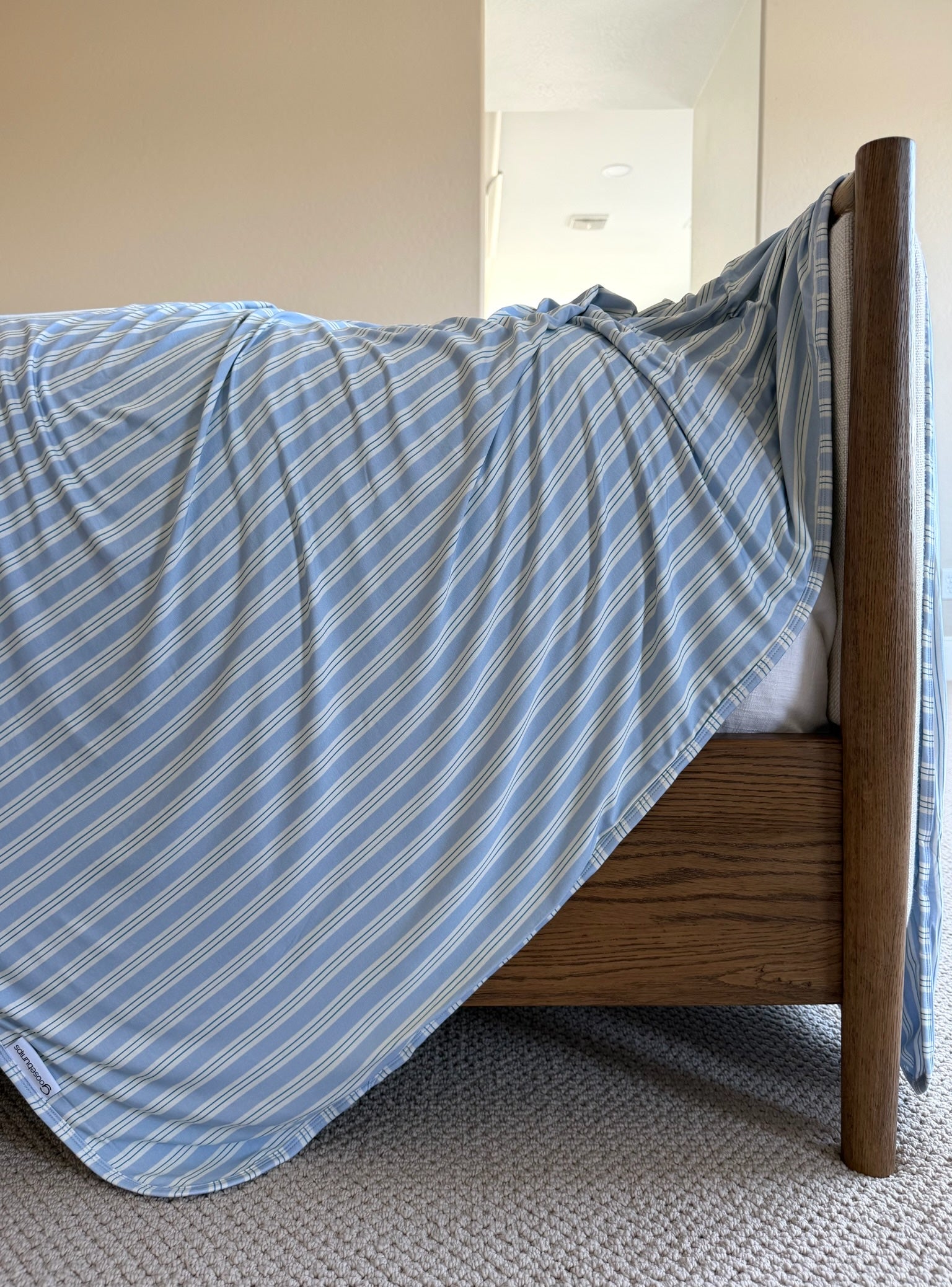 A haven wooden bed with a blue and white striped bamboo blanket draped over its side sits in a room with beige walls and carpet, with a mirror visible in the background.