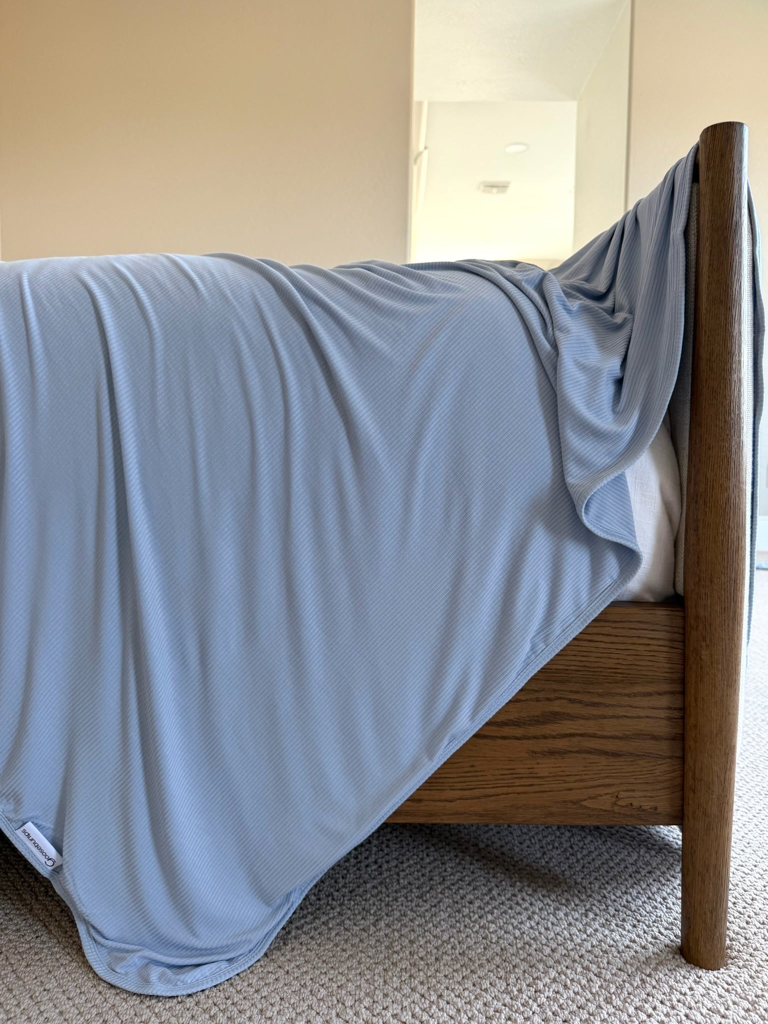A luxurious cove light blue blanket drapes over the side of a wooden bedframe in a carpeted bedroom with neutral-colored walls and a mirror in the background.