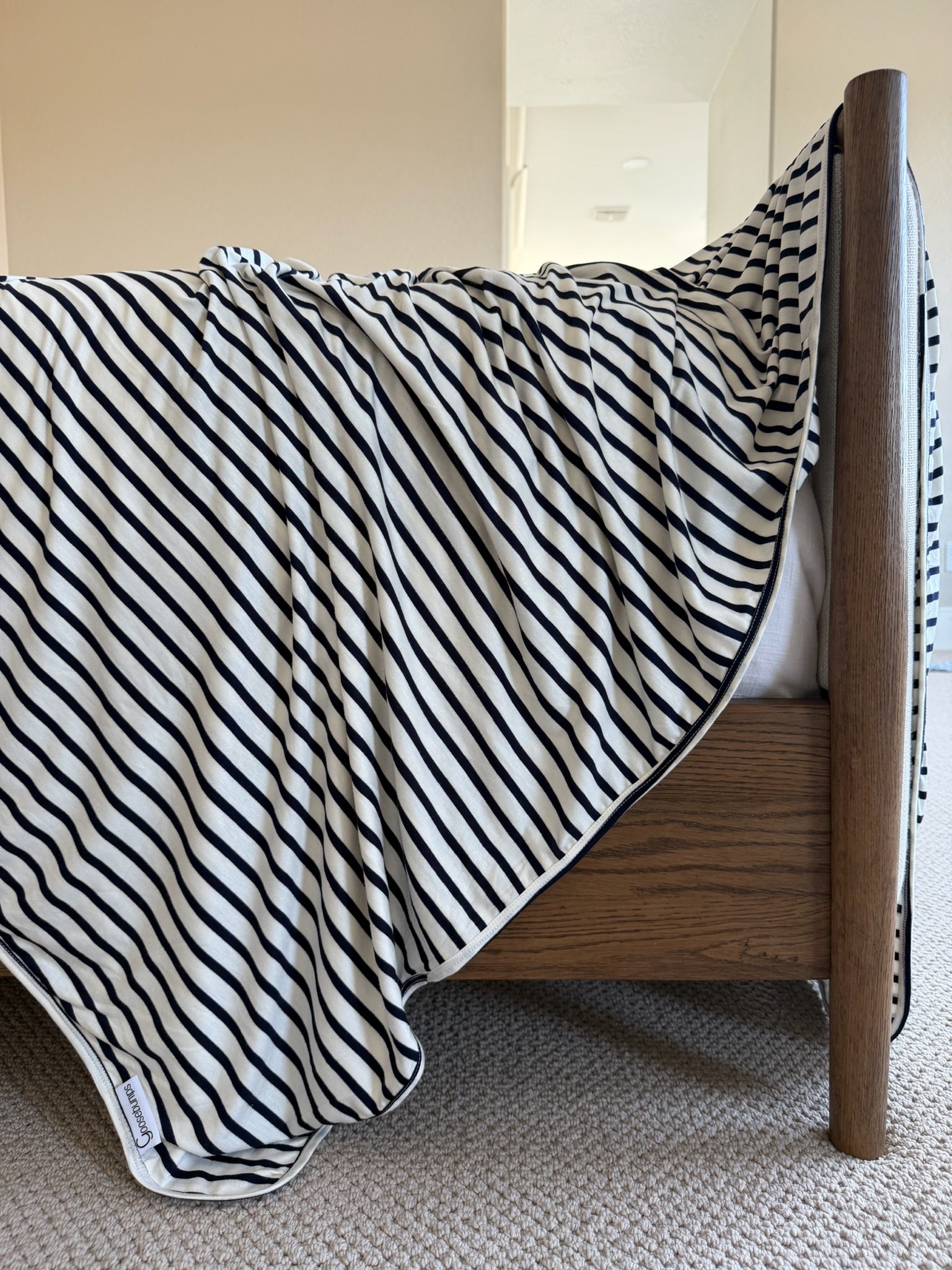 The bayside blanket, featuring luxurious black and white stripes in soft modal rayon, is casually draped over the edge of a wooden bed frame, complemented by a beige carpet and light-colored walls in the background.