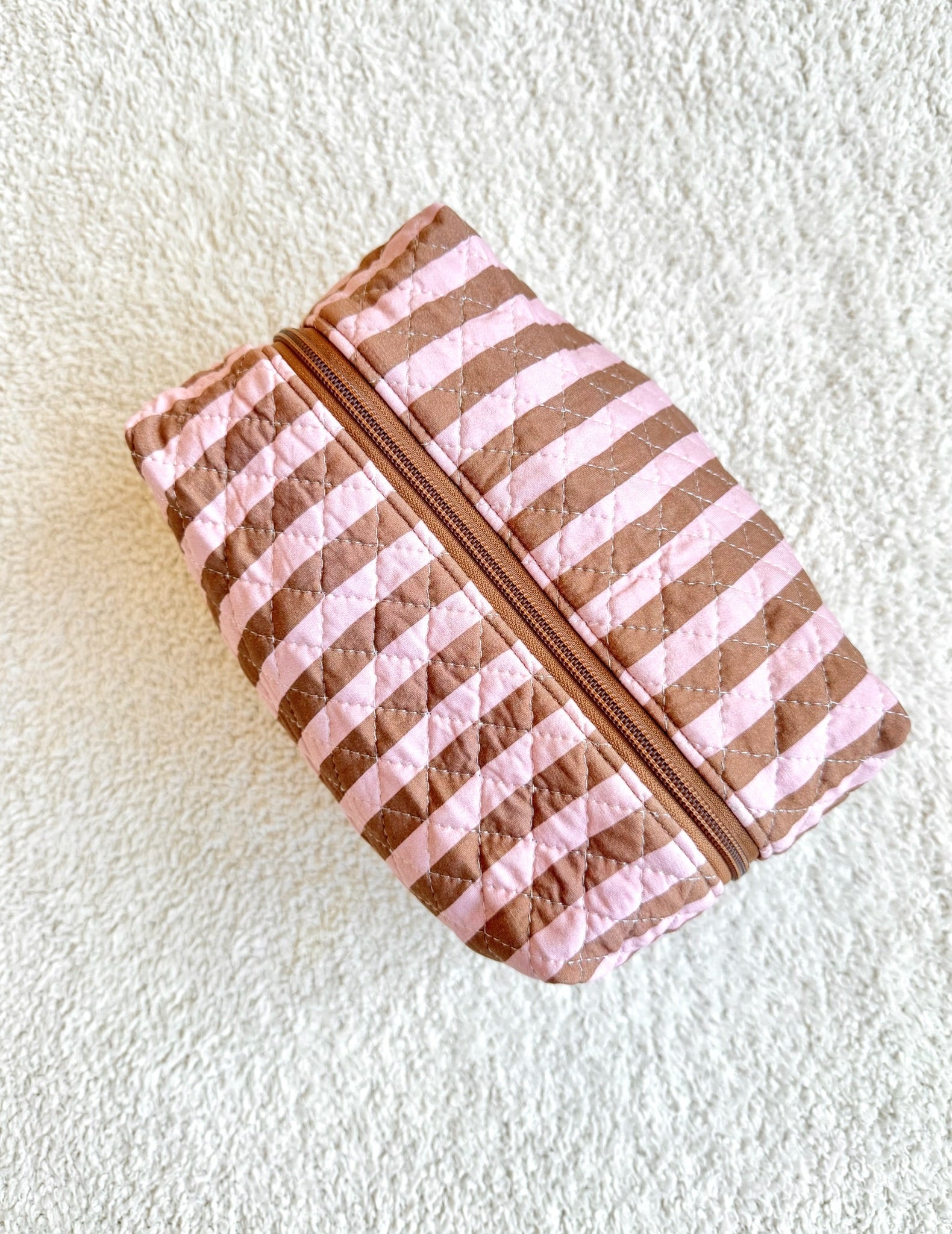 An annabelle travel bag with pink and brown diagonal quilted stripes rests on a textured white surface. Designed to elevate your travel accessories, this pouch features a central zipper for secure storage while you’re on the go.