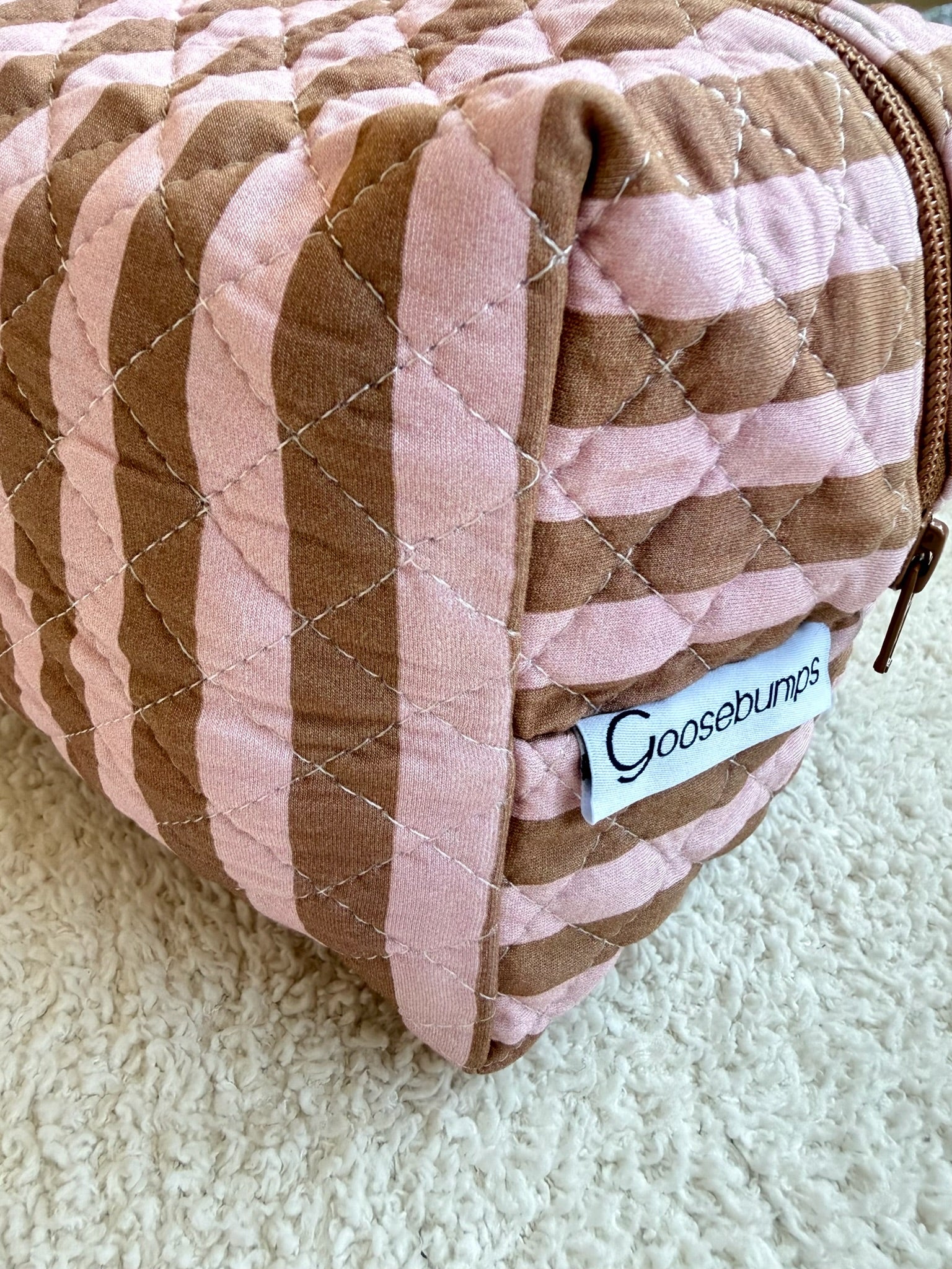 The annabelle travel bag, featuring brown and pink wavy stripes, quilted detailing, a zipper closure, and a white Goosebumps label, rests on a textured cream-colored surface—making it the perfect addition to your collection of travel accessories.