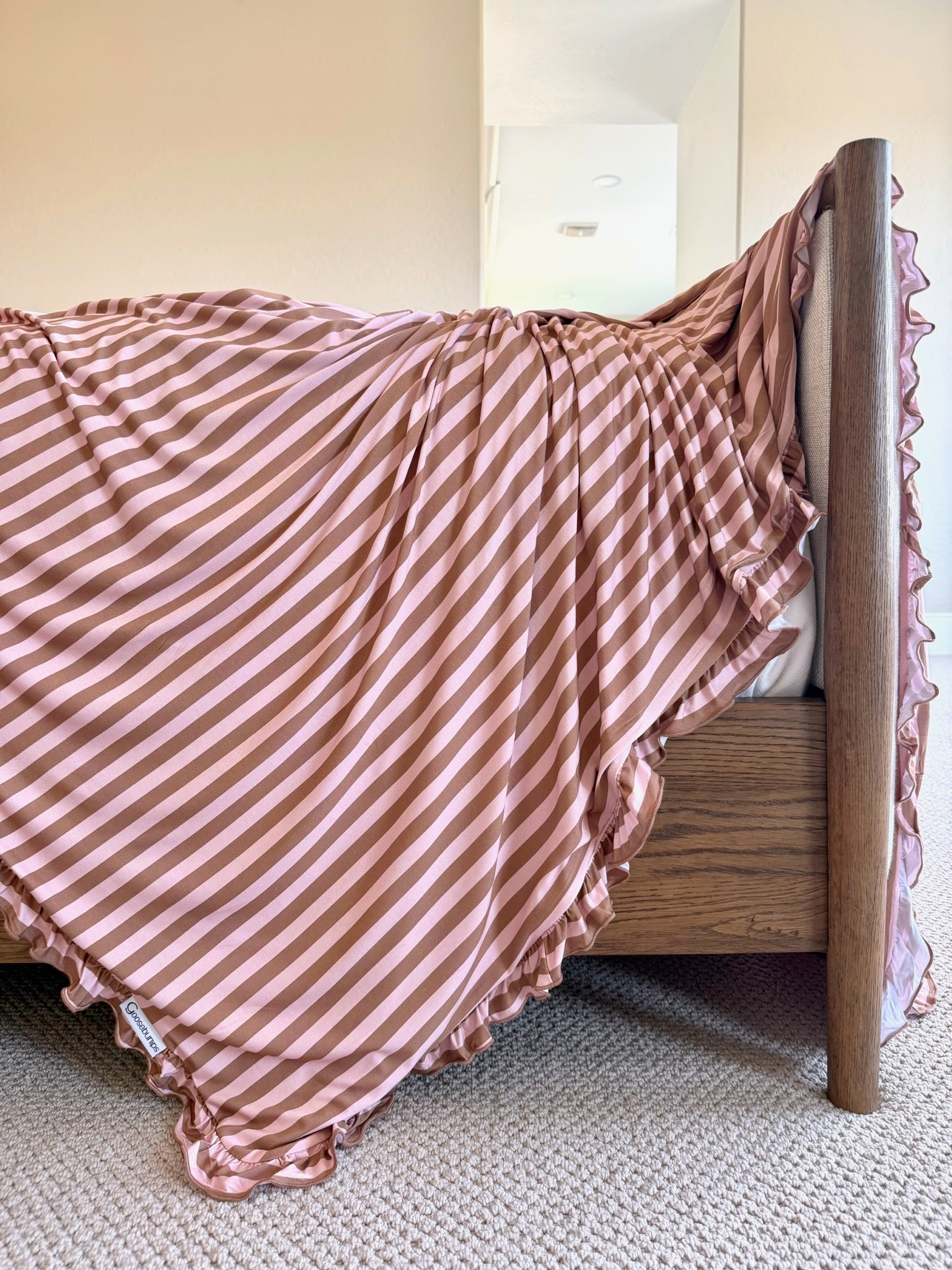 An annabelle / ruffle blanket, featuring pink and white stripes and elegant ruffled edges, is draped over the side of a wooden bed. The space has light walls, a beige carpet, and gentle sunlight streaming in from the background.