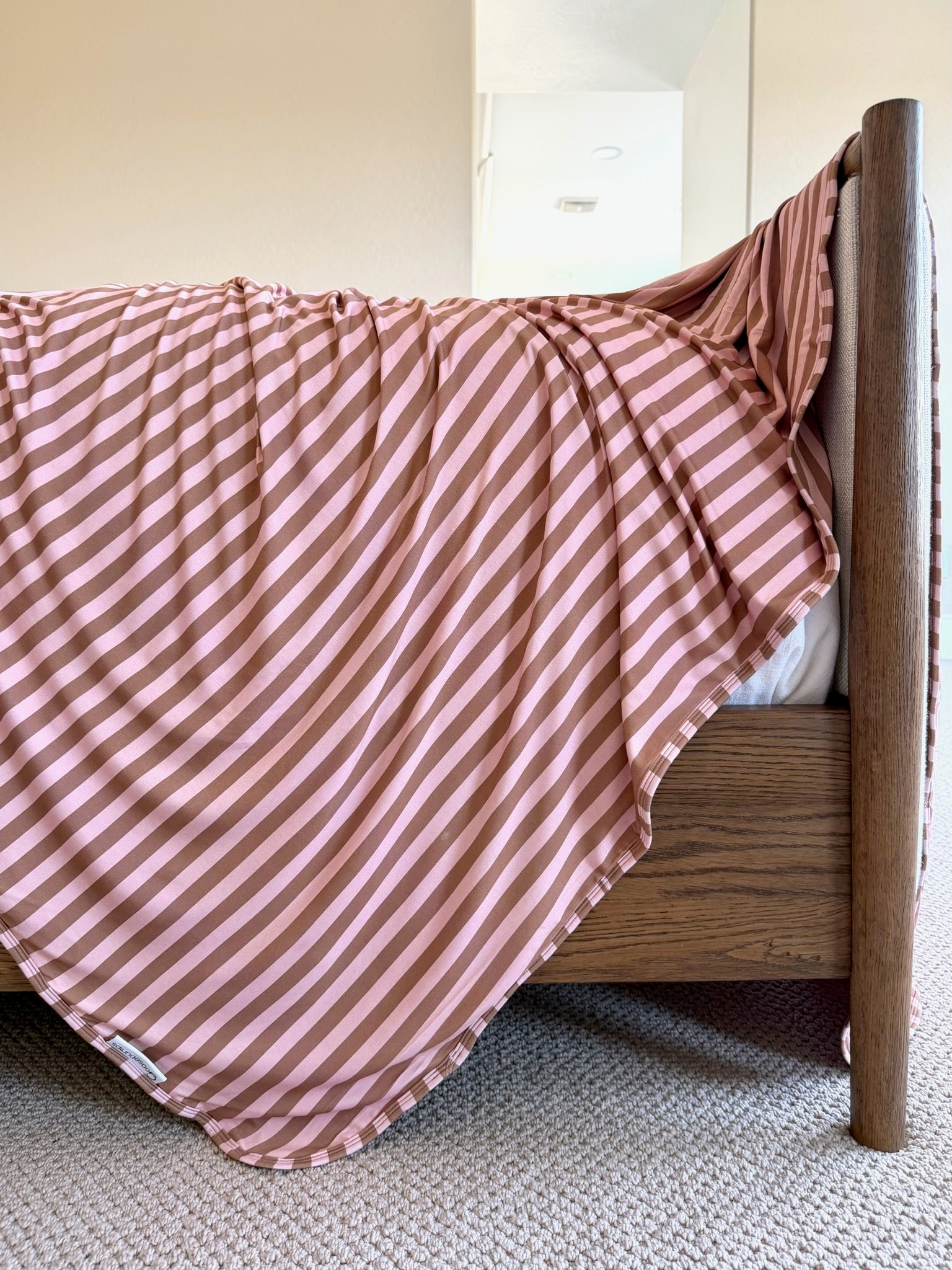 The annabelle blanket, featuring modern pink and brown stripes, is draped over the side of a wooden bed frame in a sunlit, carpeted bedroom.