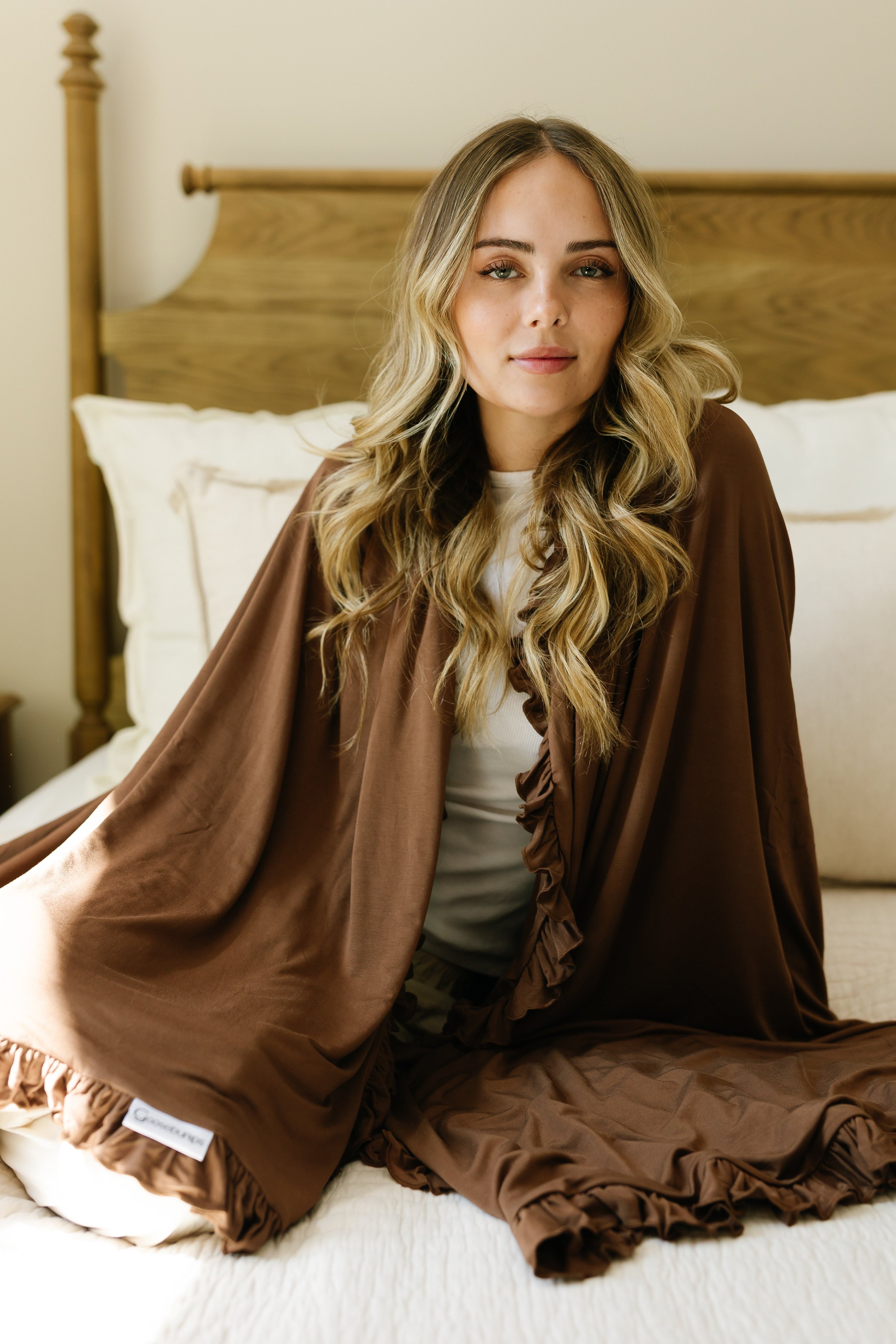 A woman with long, wavy blonde hair sits on a bed, wrapped in the chocolate / ruffle blanket. She looks at the camera as soft natural light fills the room.