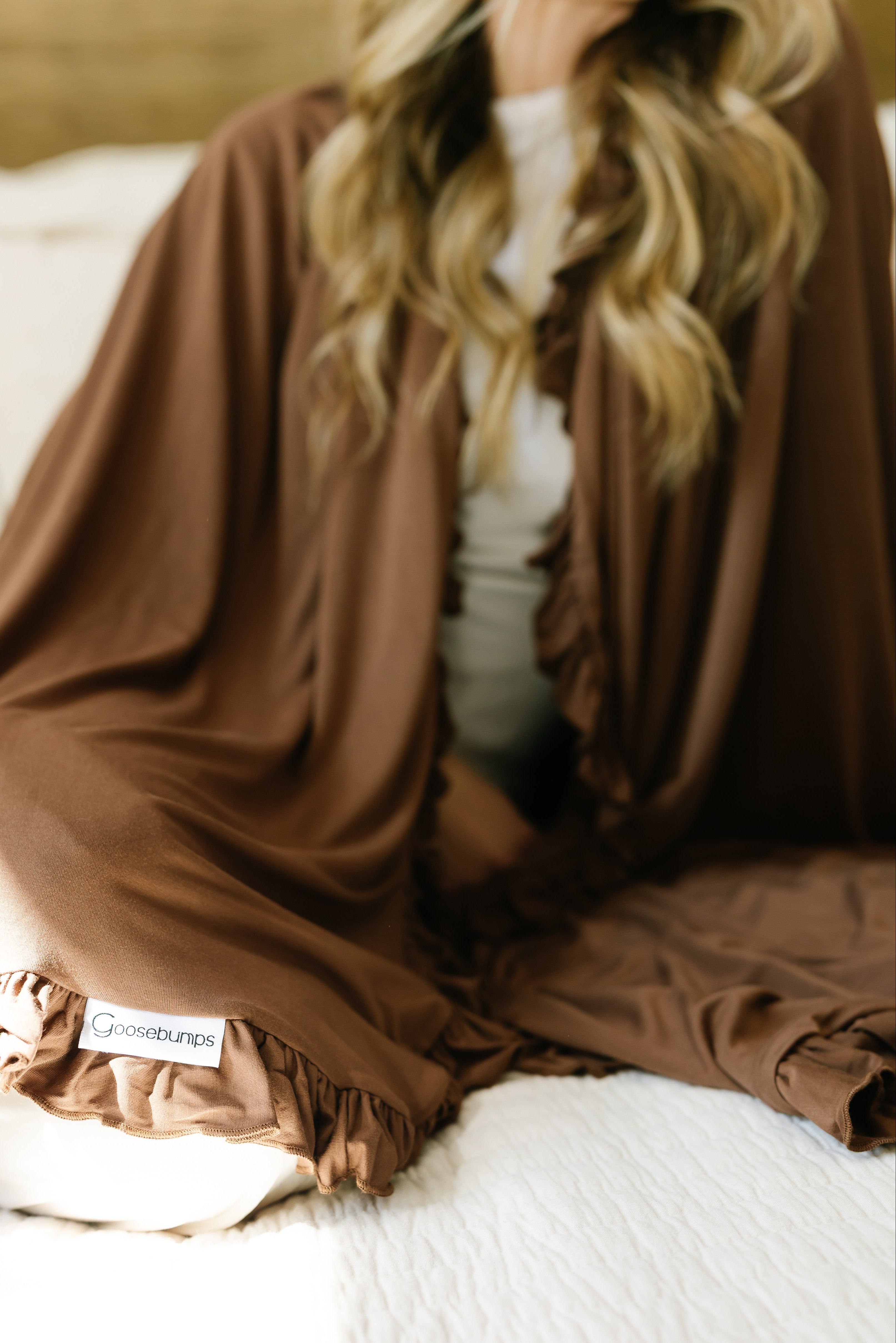 A woman with long, wavy hair sits on a bed wrapped in a chocolate ruffle blanket. The cozy, relaxed setting is bathed in soft natural light.
