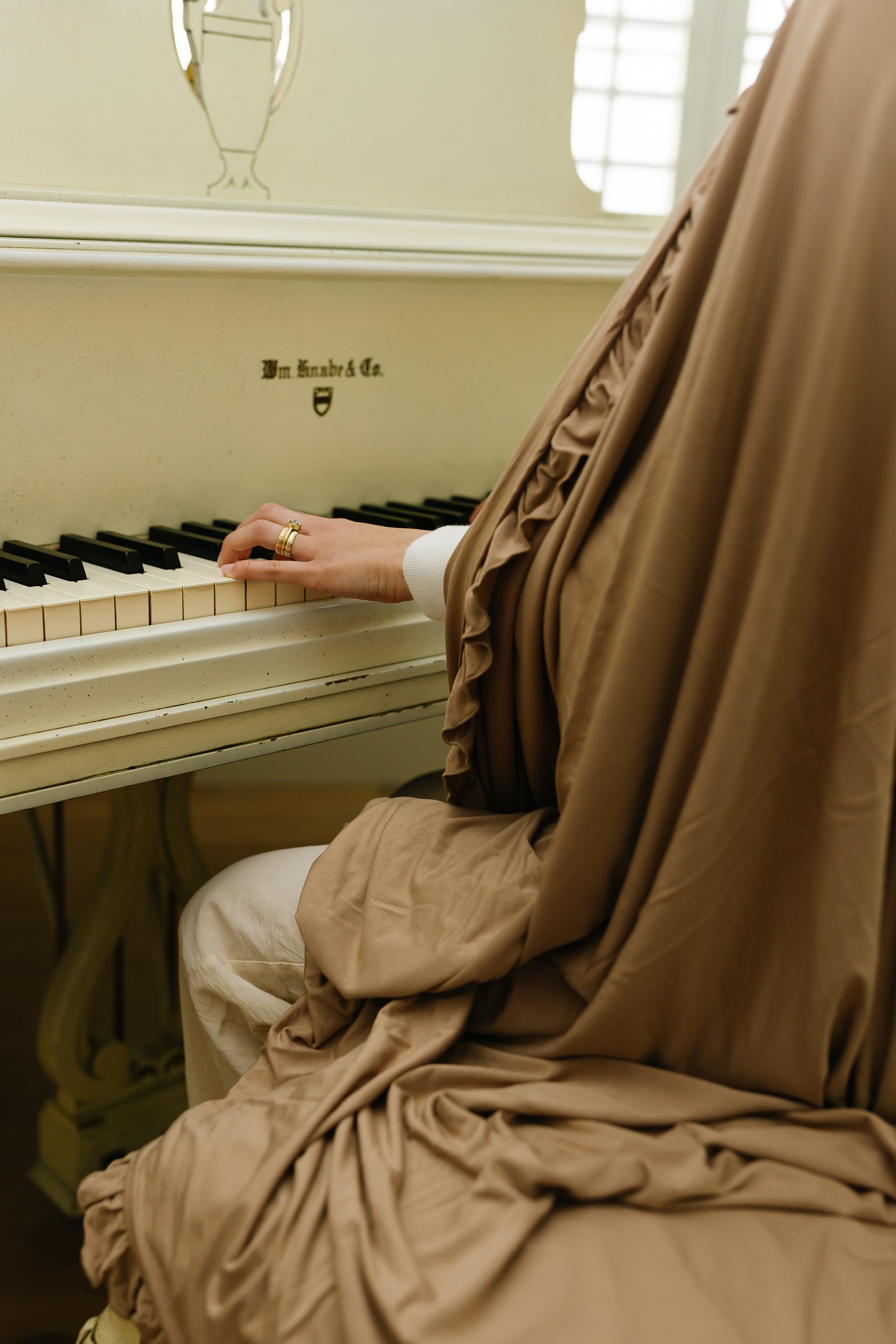 A hand wearing a gingerbread / ruffle bamboo blanket plays a white piano, highlighting the piano keys and a gold ring, with only part of the person’s body and hand visible.